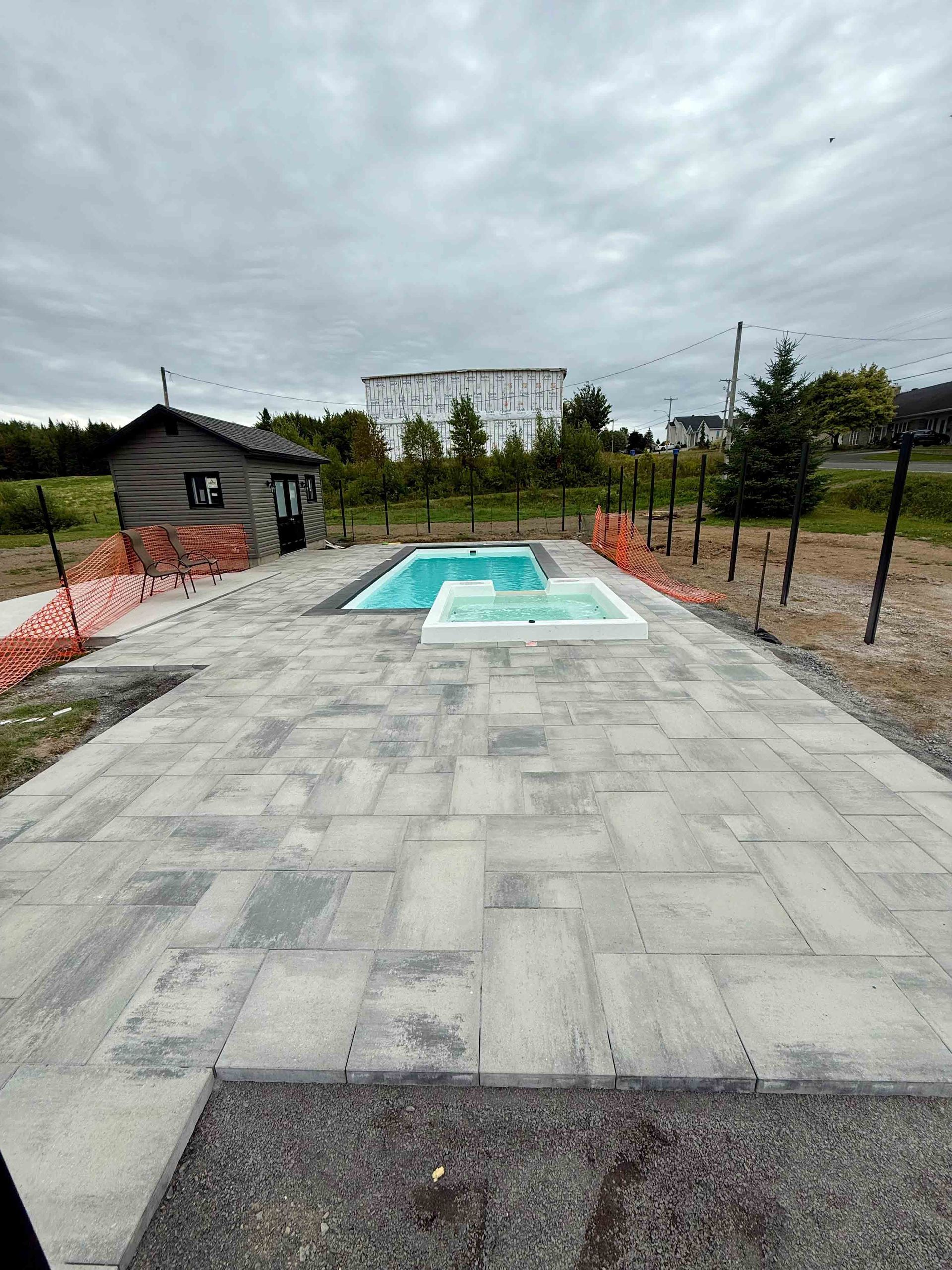 Allée en béton menant à une petite piscine turquoise dans une cour extérieure clôturée sous un ciel nuageux