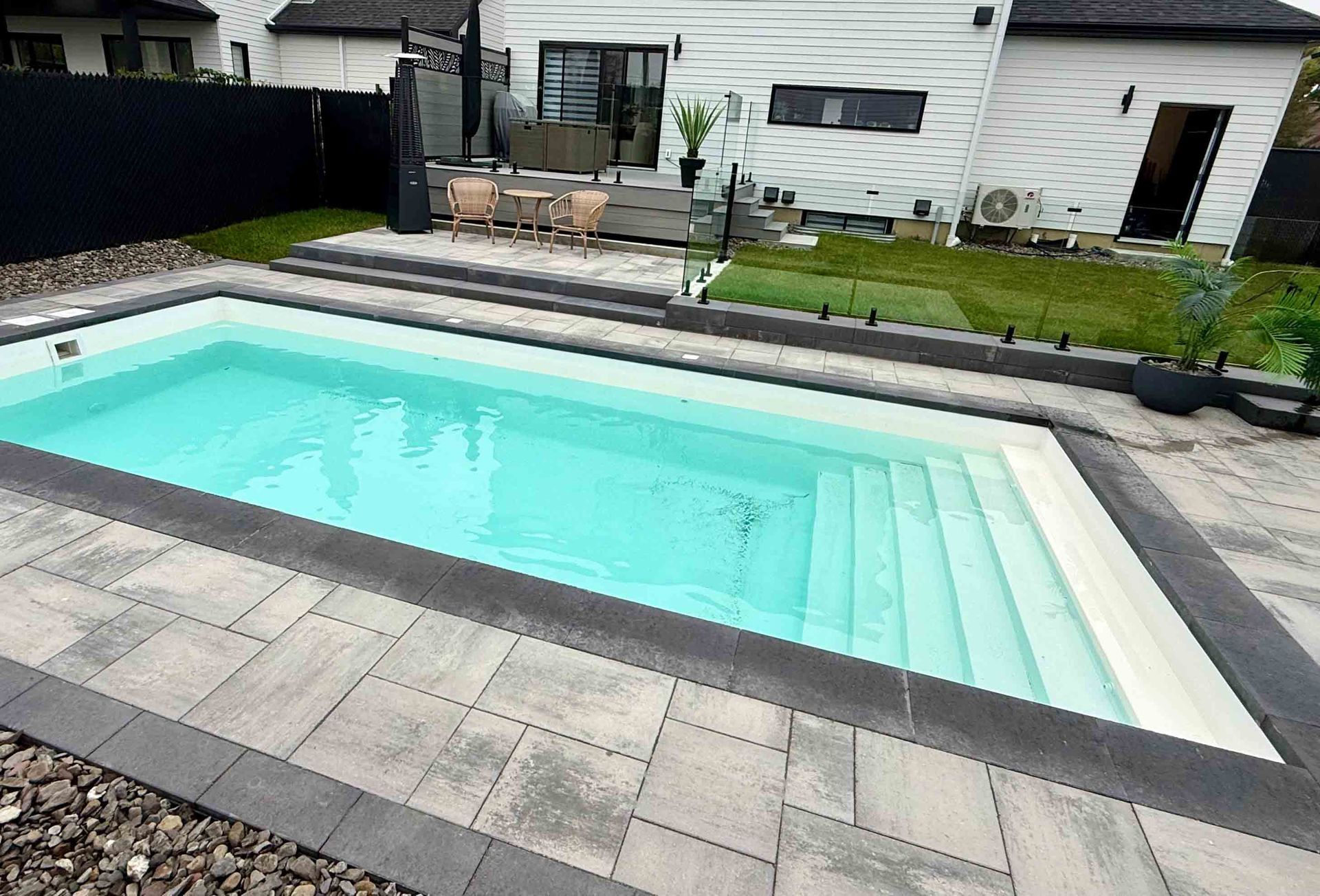 Piscine rectangulaire dans un jardin avec une eau bleu clair, une terrasse en pierre et une maison blanche en arrière-plan.