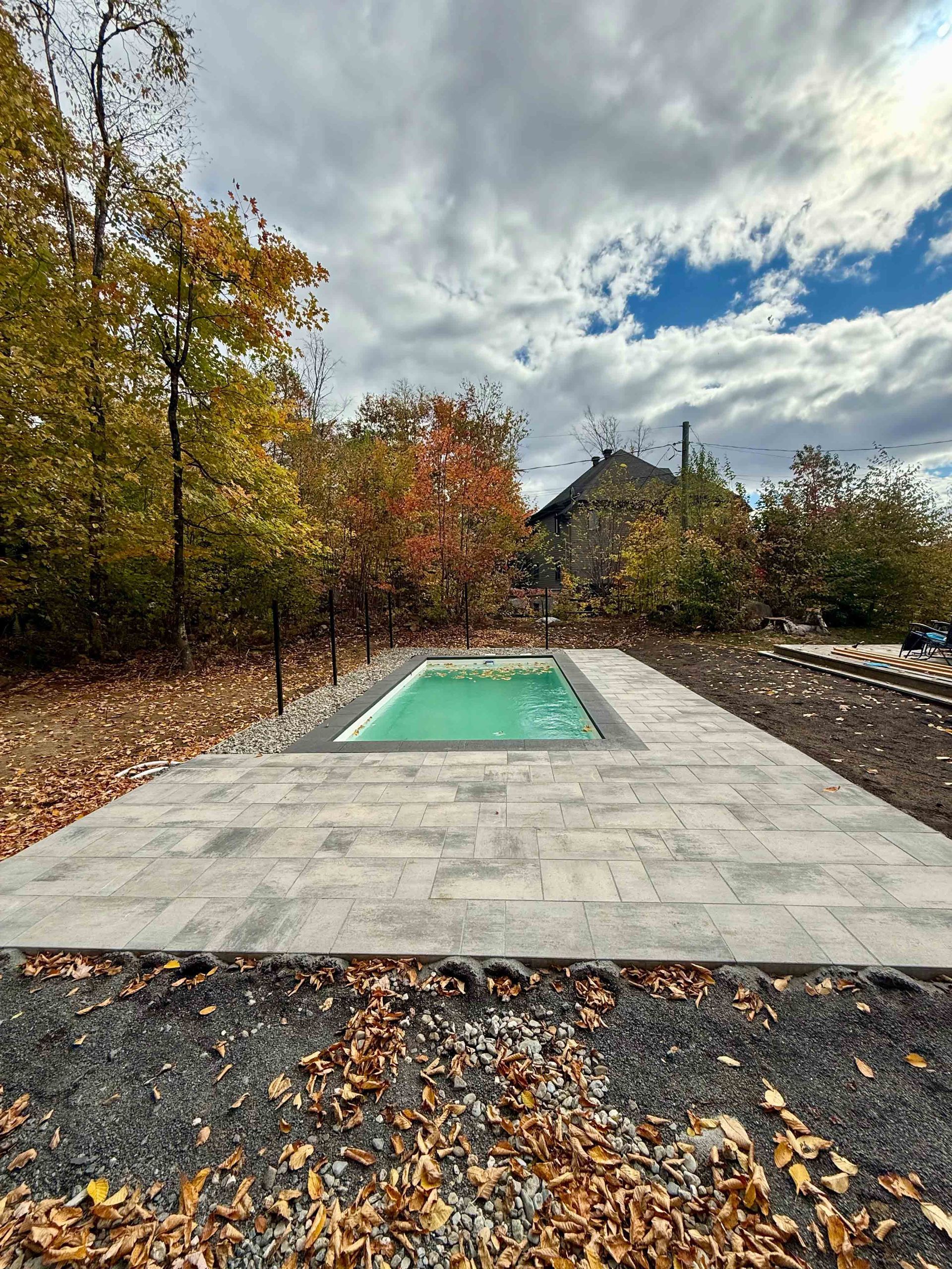 Terrasse extérieure avec une petite piscine rectangulaire verte, entourée d'arbres d'automne et d'un ciel nuageux