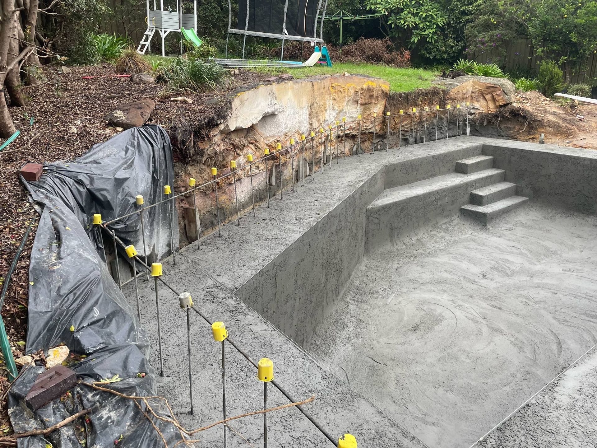 Excavated backyard pool under construction, with concrete steps and black tarp along the left wall.