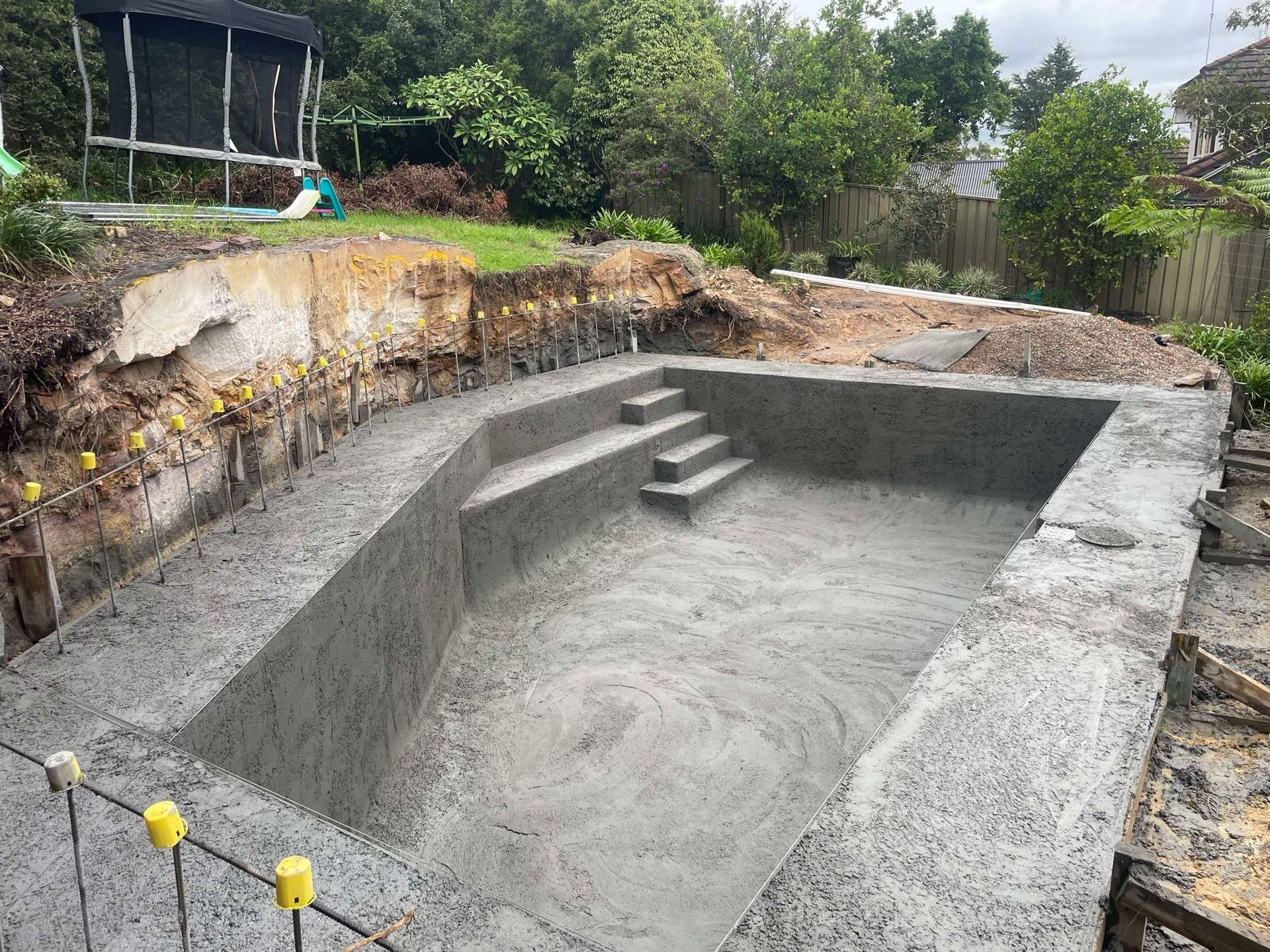 Empty concrete swimming pool under construction in a backyard, with steps and surrounding dirt excavation.