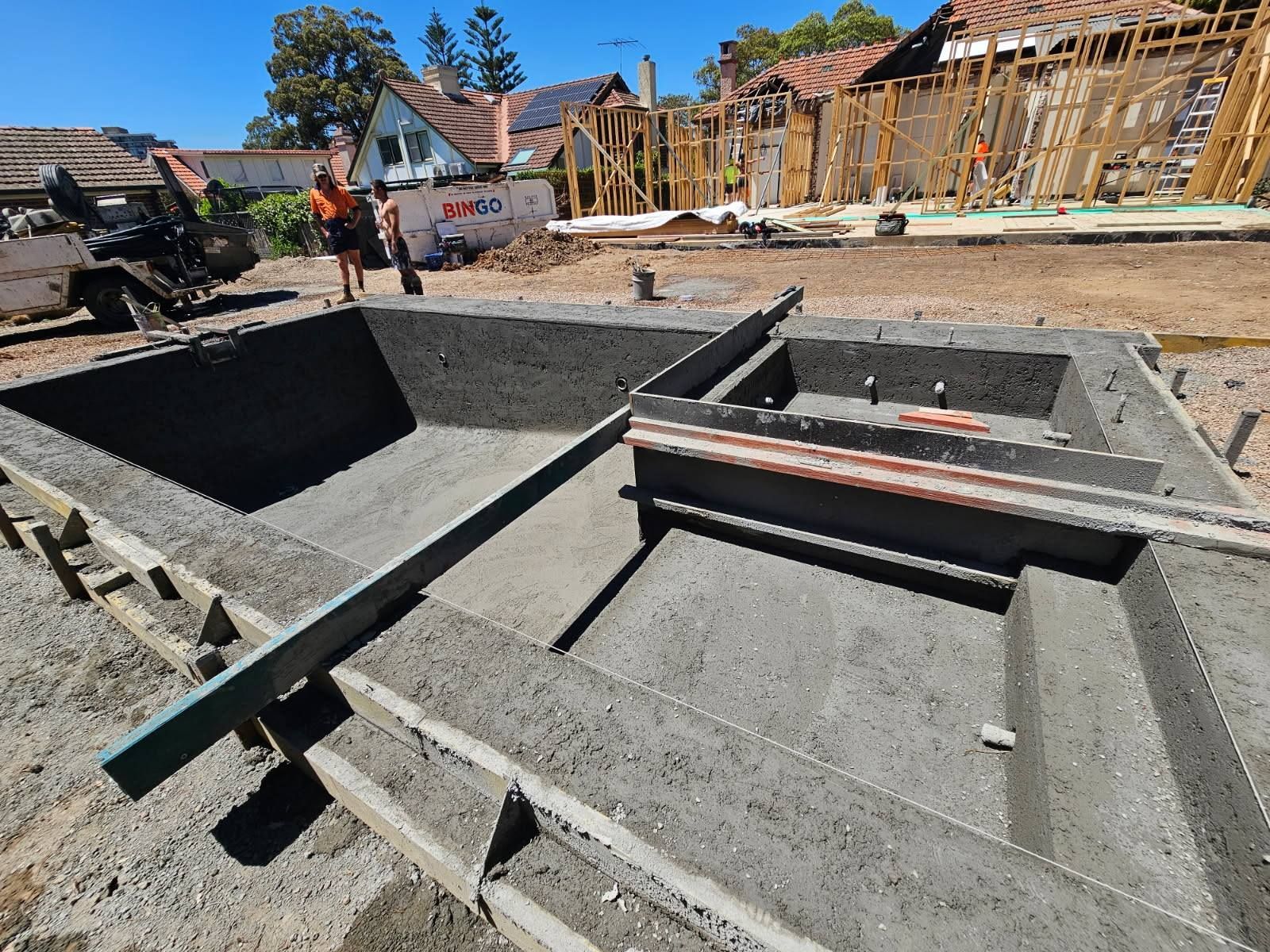 Concrete foundation and framing at a house construction site with workers and wooden scaffolding.