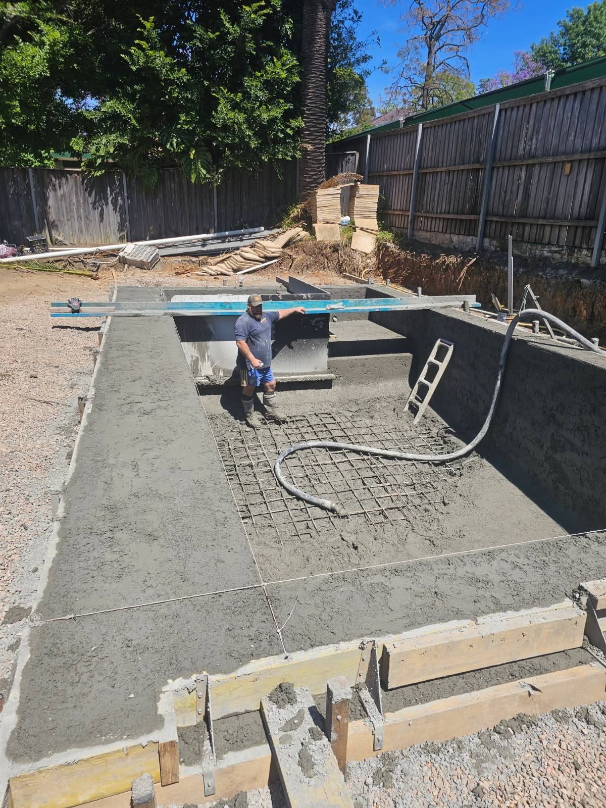 Backyard swimming pool under construction, with a worker standing in a concrete excavation.