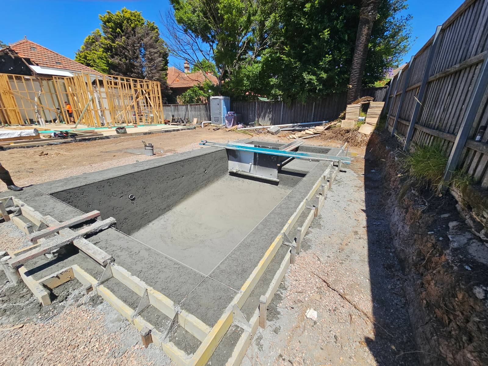 Partially built backyard in-ground pool under construction beside a wooden fence and trees.