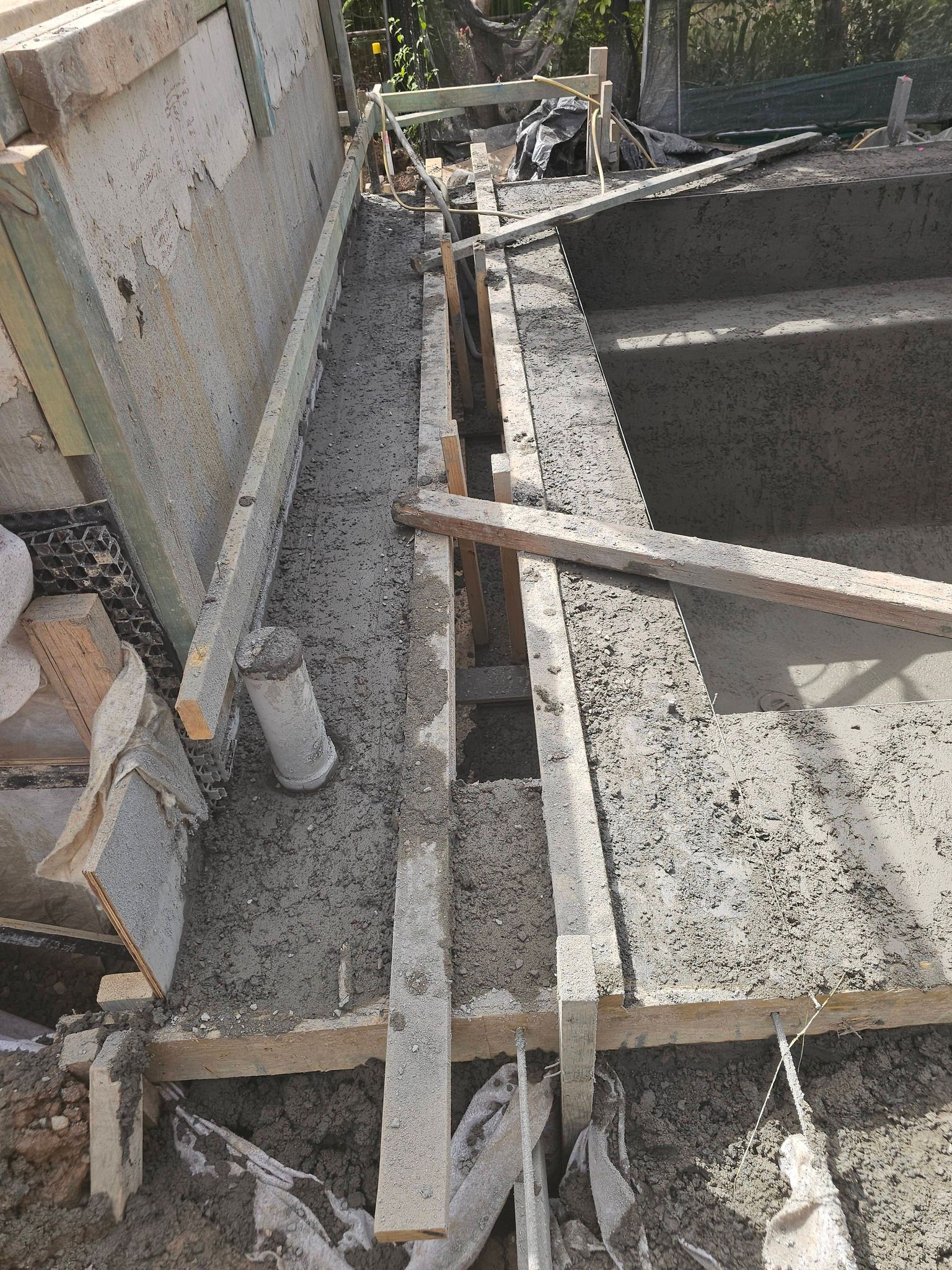 Construction site with wet concrete, wooden formwork, and a rebar framework beside a wall.