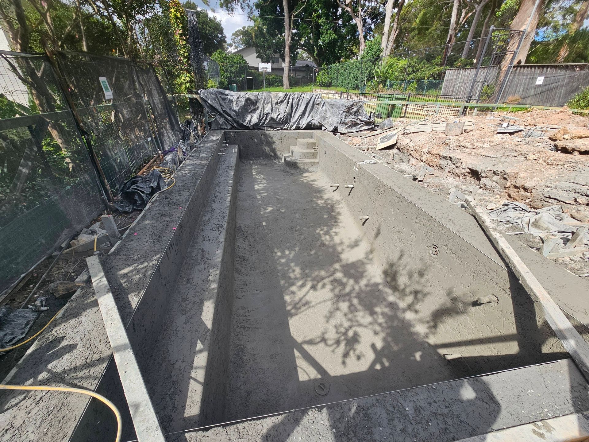Empty concrete trench under construction beside a fenced, wooded area with debris around it