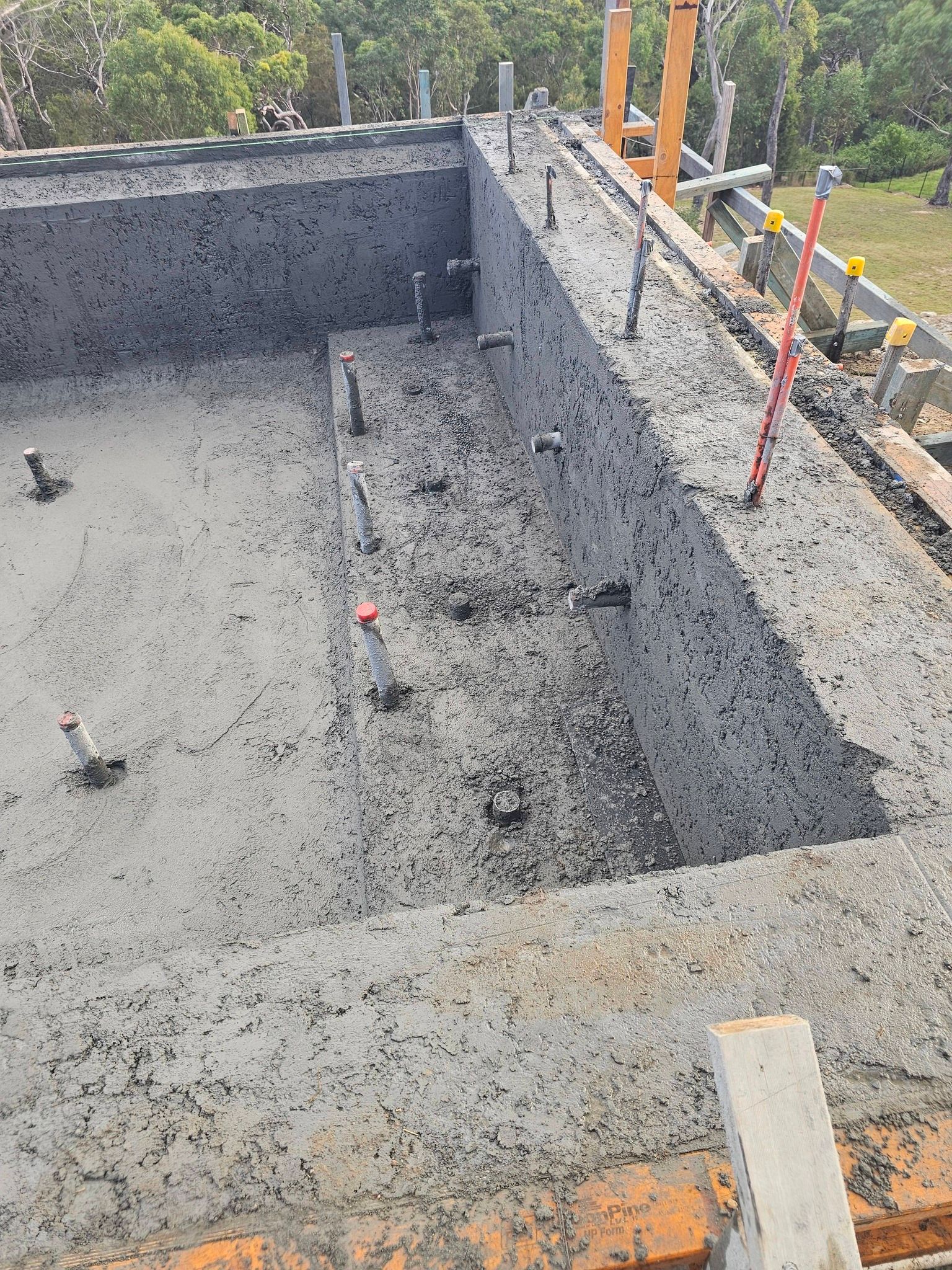 Concrete foundation pit at a construction site, with gravel walls, rebar, and orange safety poles.