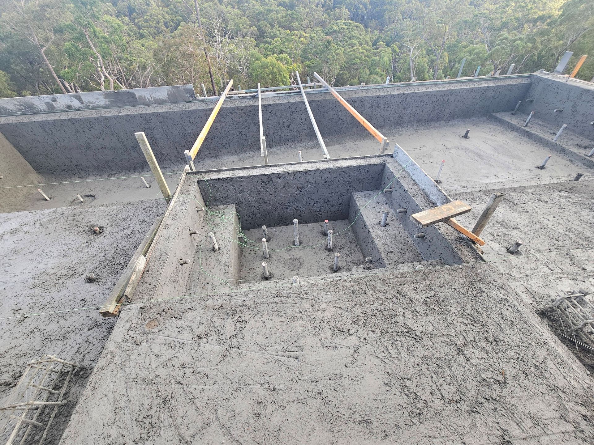 Concrete rooftop construction with a central opening, rebar, and forested hillside in the background