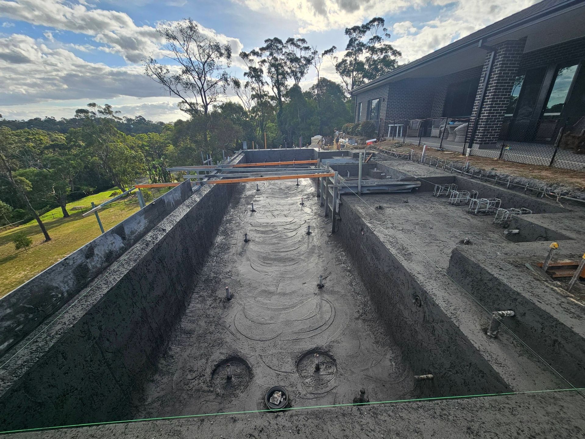 Concrete pool under construction beside a hillside building, with a wet foundation and excavation equipment.