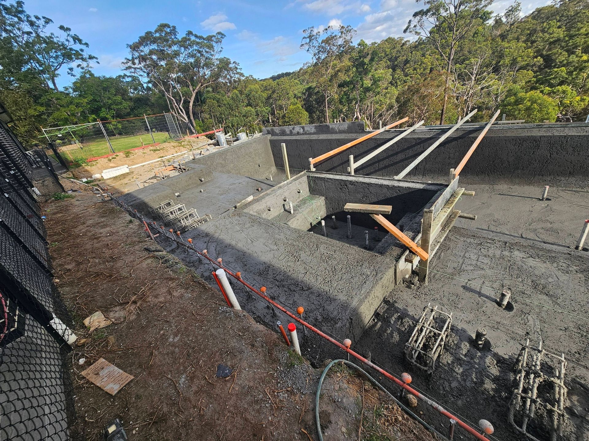 Cement-lined excavation with wooden braces and orange safety netting on a hillside under a blue sky
