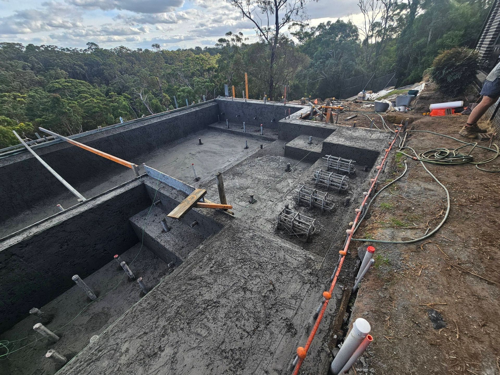 Concrete pool construction site with wet foundation, rebar, tools, and forested hillside background