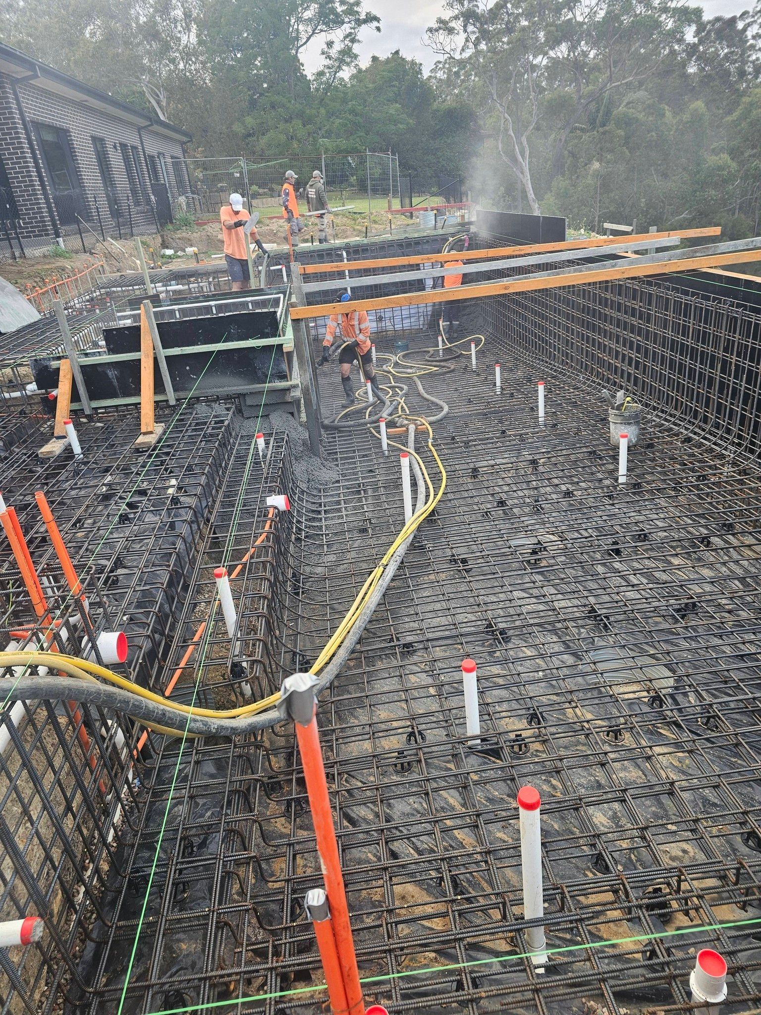 Construction excavation site with workers, orange barriers, and exposed rebar on a muddy foundation
