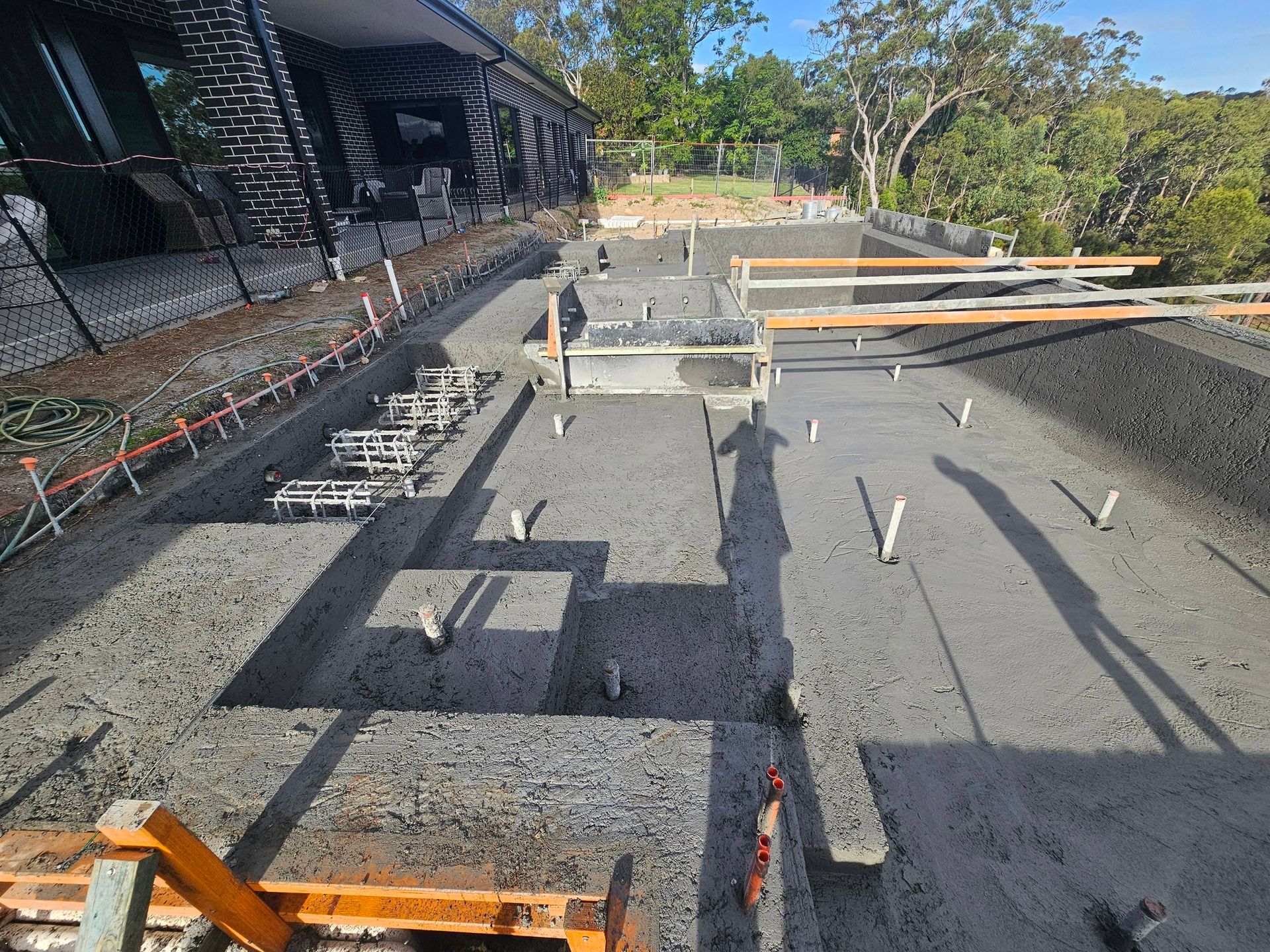 Outdoor construction site with foundation forms, rebar, and concrete slabs beside a building under a blue sky