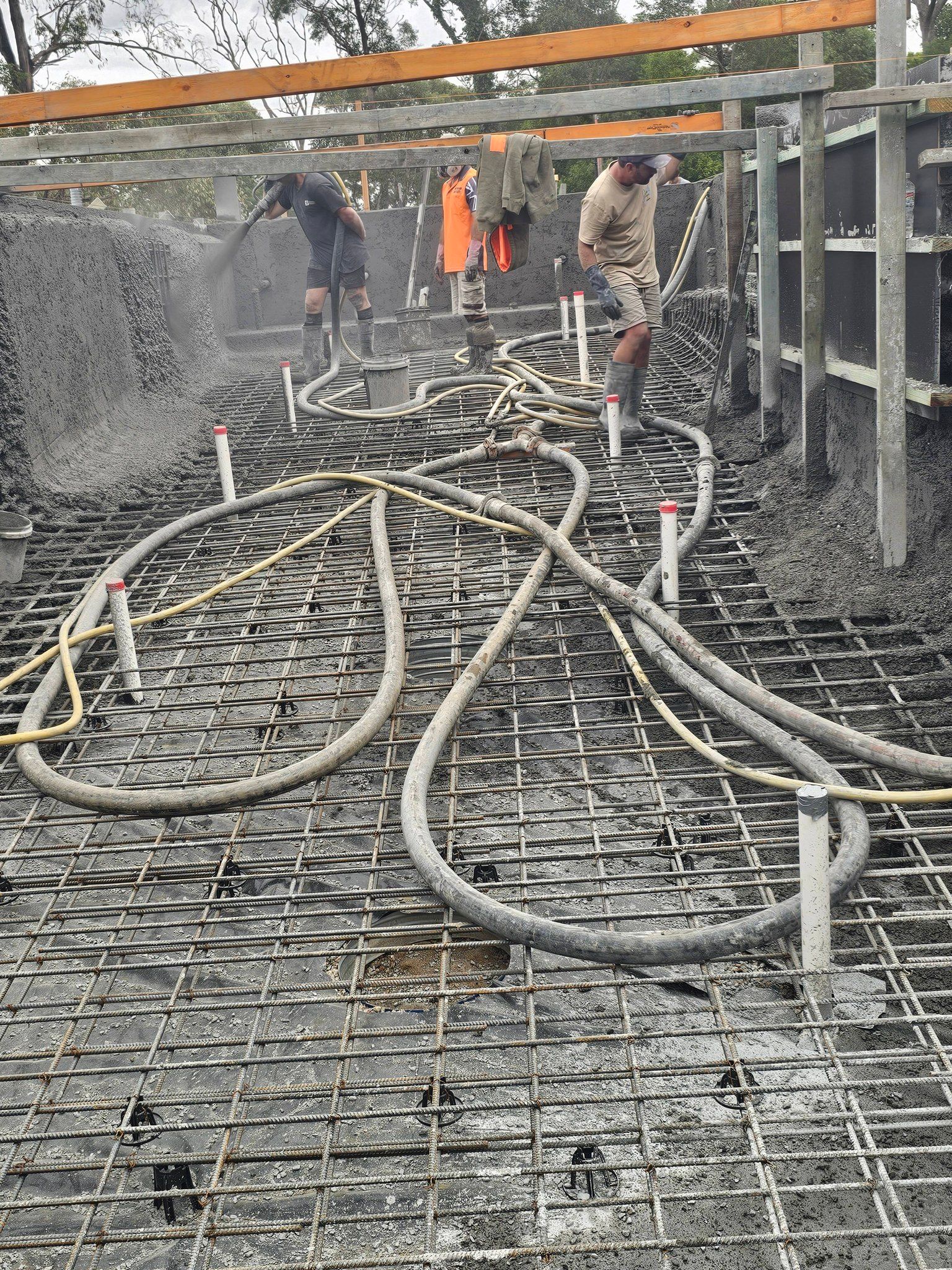 Construction workers laying rebar and concrete hoses on a wet foundation slab under steel framing.