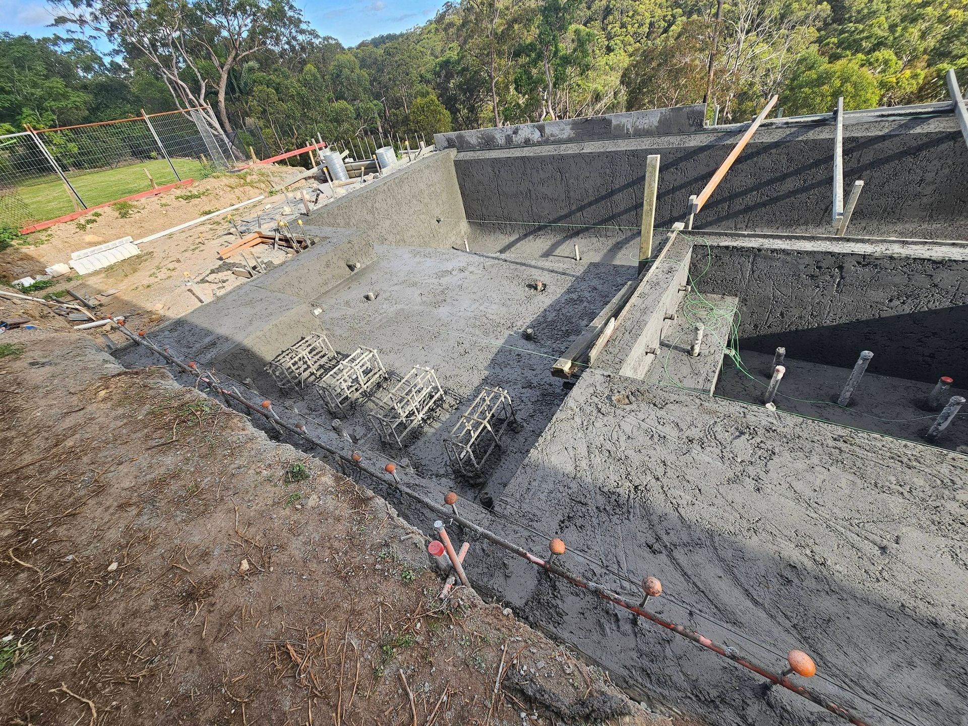 Fresh concrete foundation walls and slabs under construction at a wooded site, with wooden braces and forms.