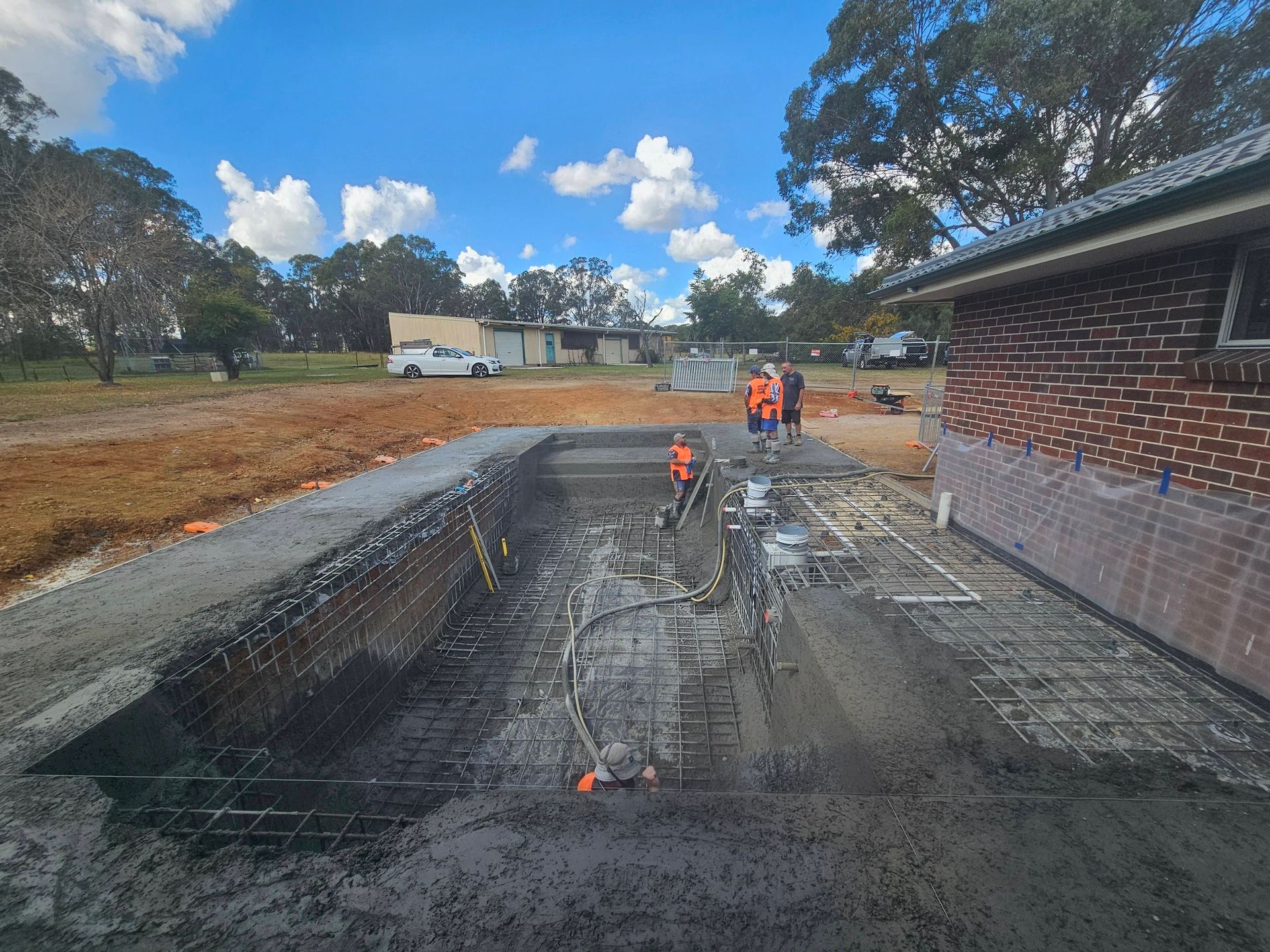 Workers installing rebar in a concrete foundation trench beside a brick house under a blue sky