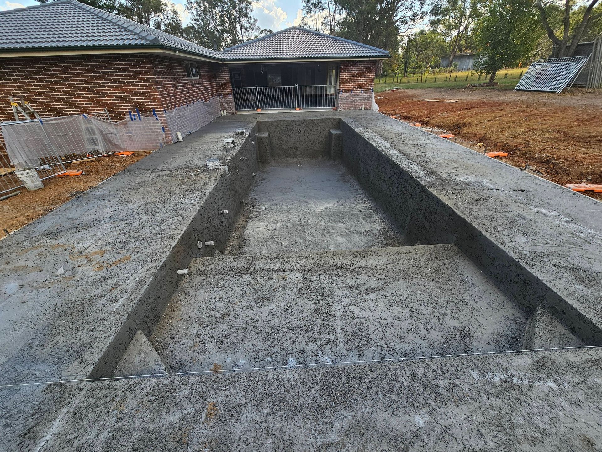 Concrete rectangular pool foundation beside a house under construction, with a gravel yard and roof in back
