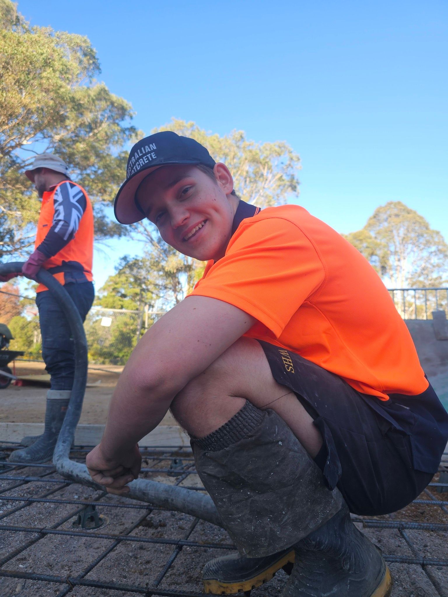 Person in an orange shirt crouches outdoors on a paved path, with another person standing in the background.