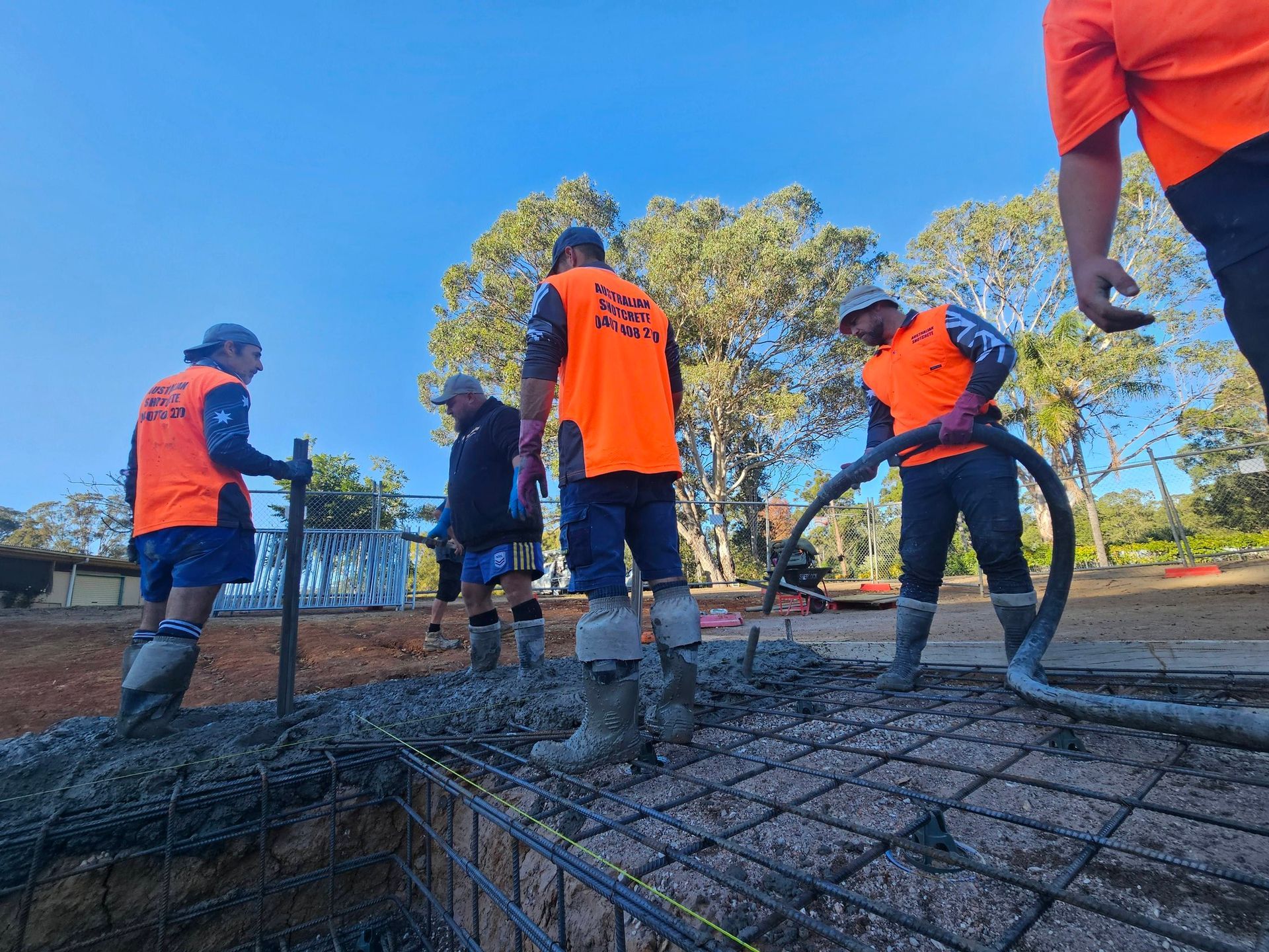 Workers in orange safety vests pour concrete over rebar at a construction site.