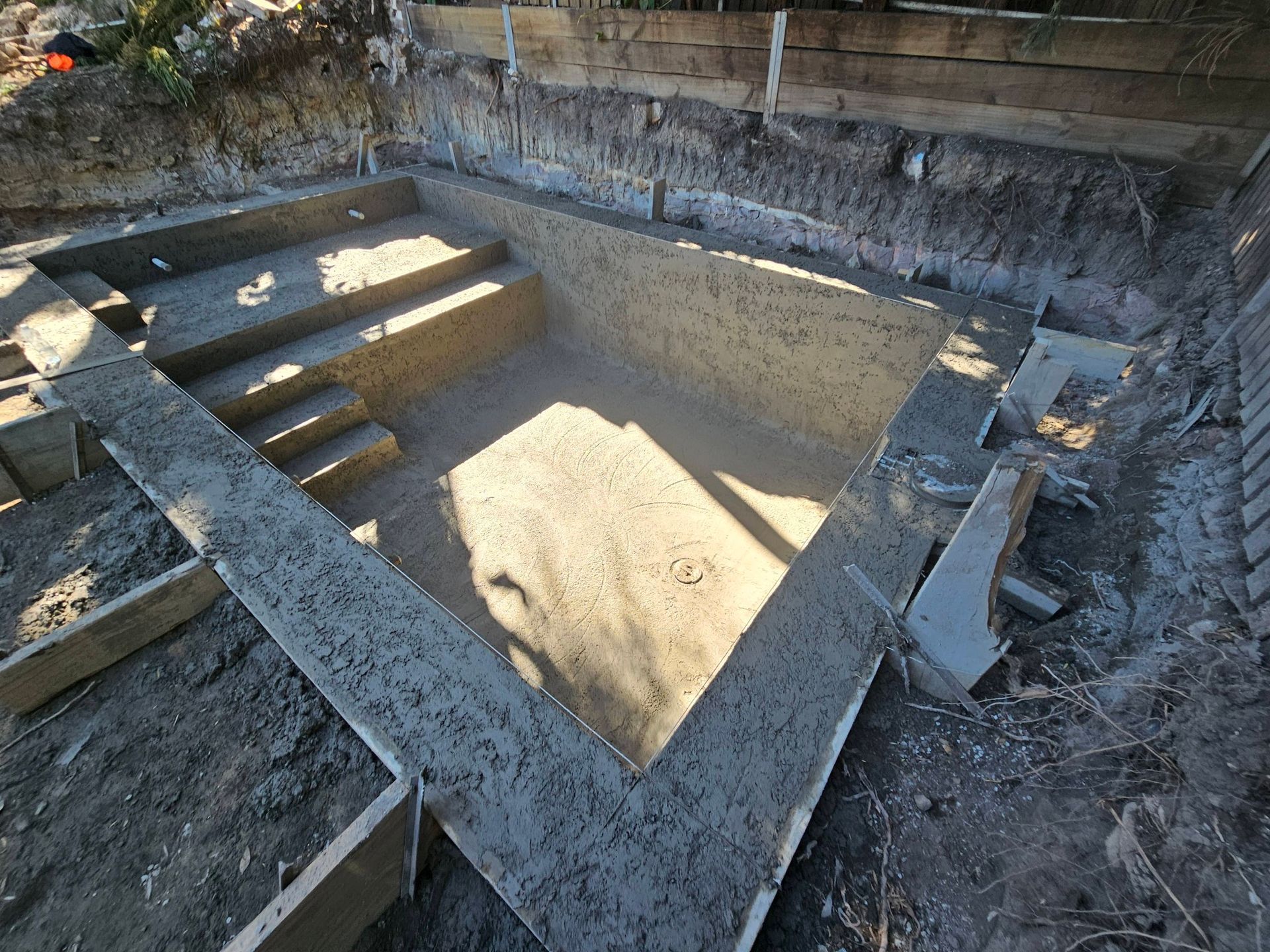 Concrete pool foundation under construction, with steps and sandy floor in a dug-out backyard area.