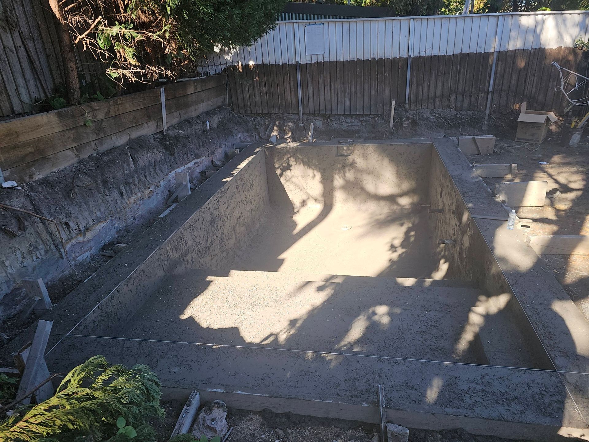 Empty concrete swimming pool in a fenced backyard, with sunlight and tree shadows.