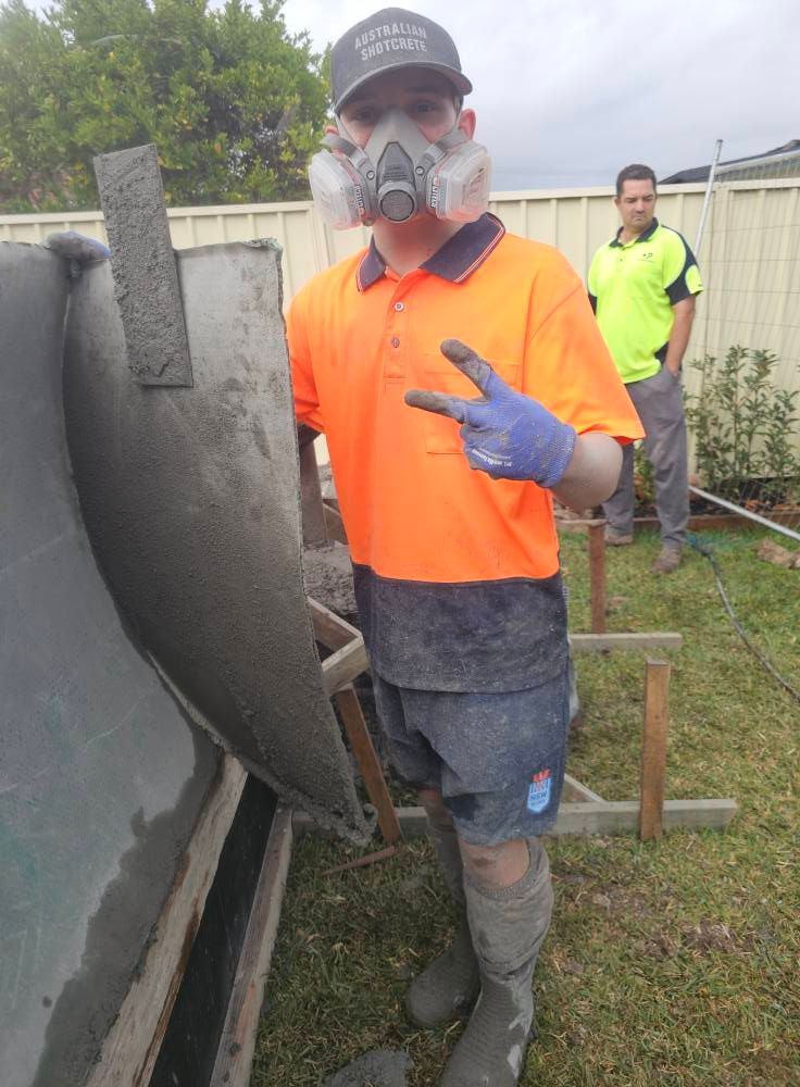 Worker in orange vest and dust mask standing by a concrete wall, with another worker in a neon shirt behind him.
