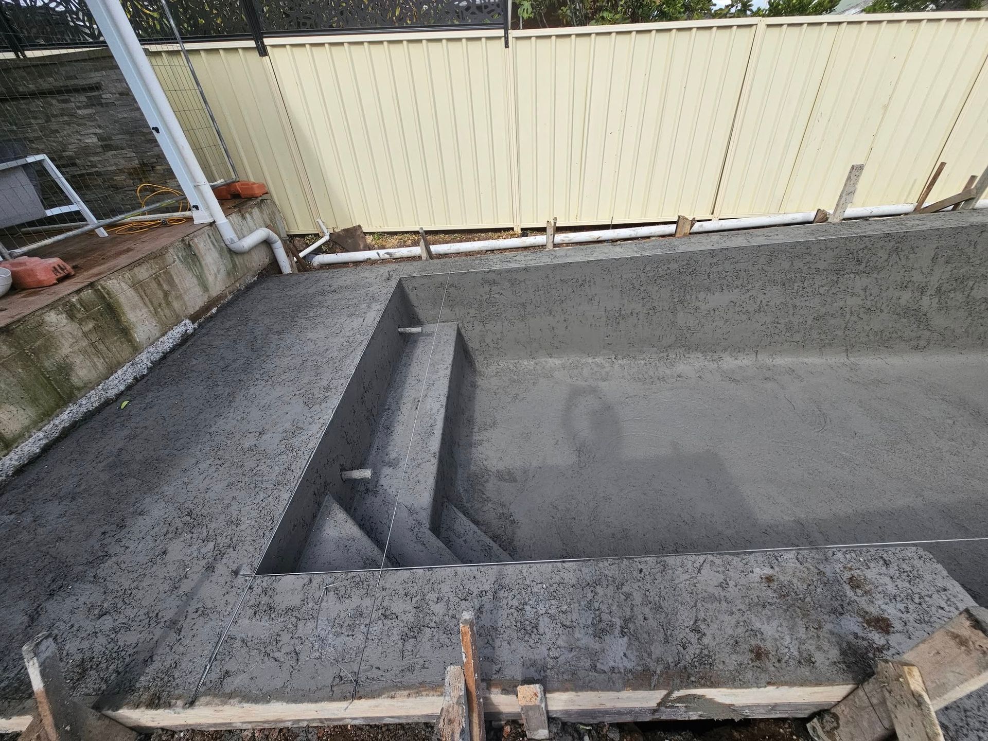 Concrete drainage basin under construction beside a beige fence in a gravel yard