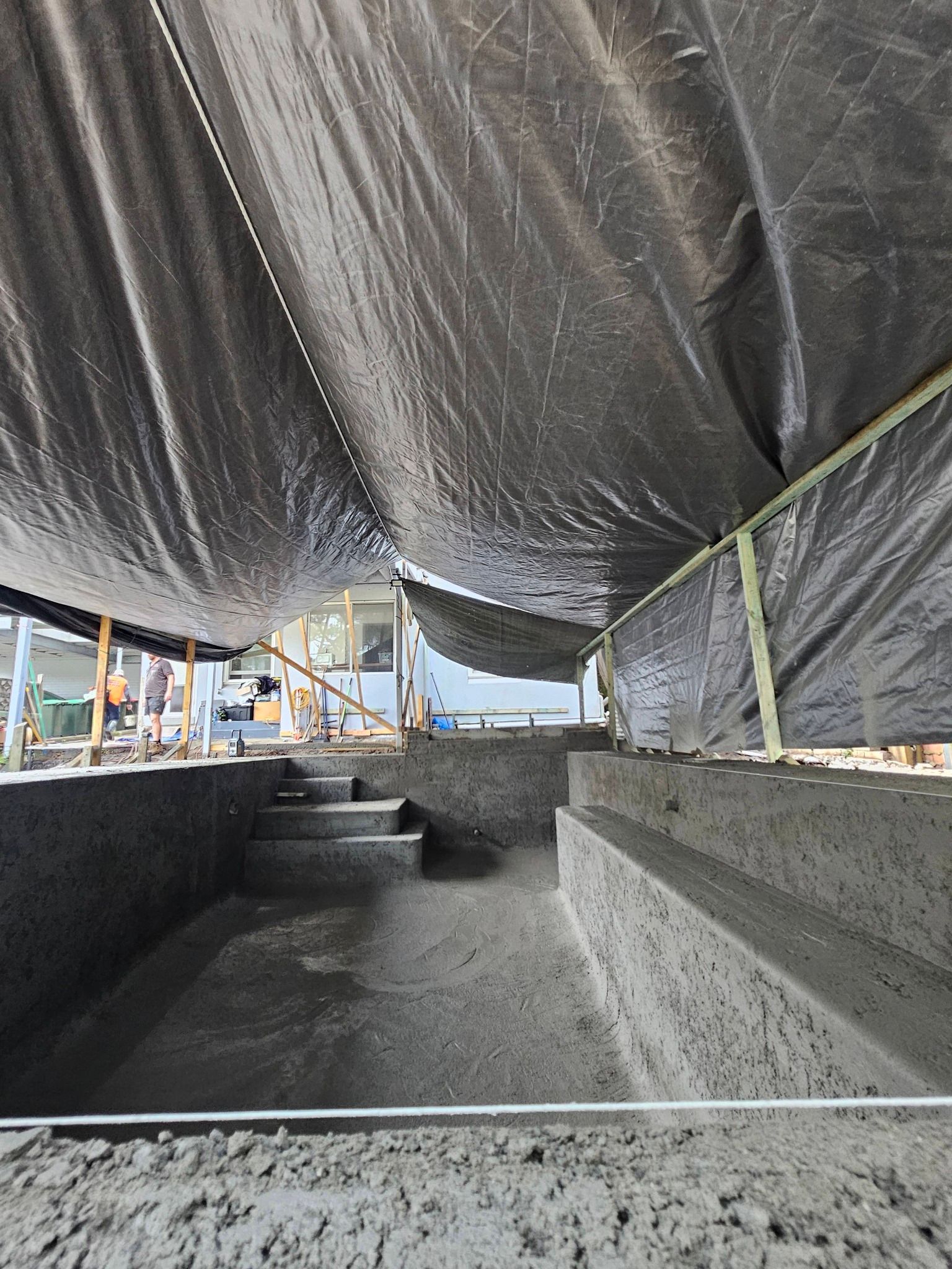 Covered concrete excavation under black tarps with steps and rebar at a construction site