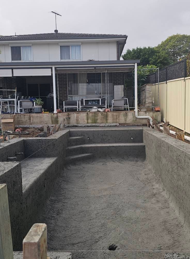 Empty concrete backyard pool under construction behind a two-story house with patio furniture.