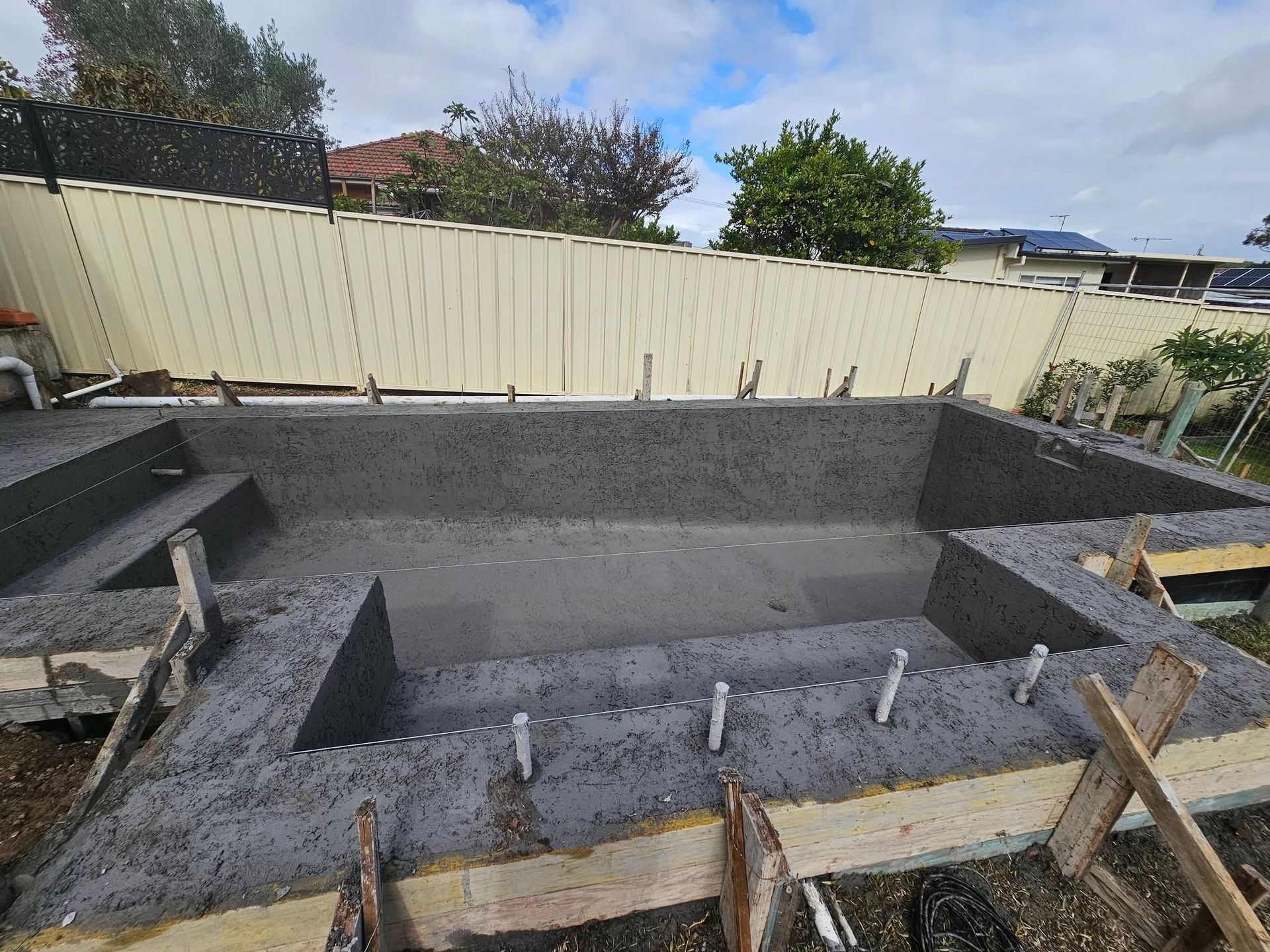 Concrete pool shell under construction in a backyard, with wooden braces and a beige fence behind it