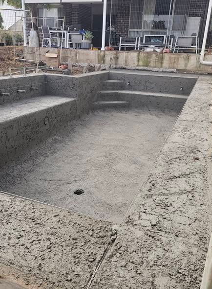 Empty concrete in-ground pool under construction, with steps and rough edges in a backyard.
