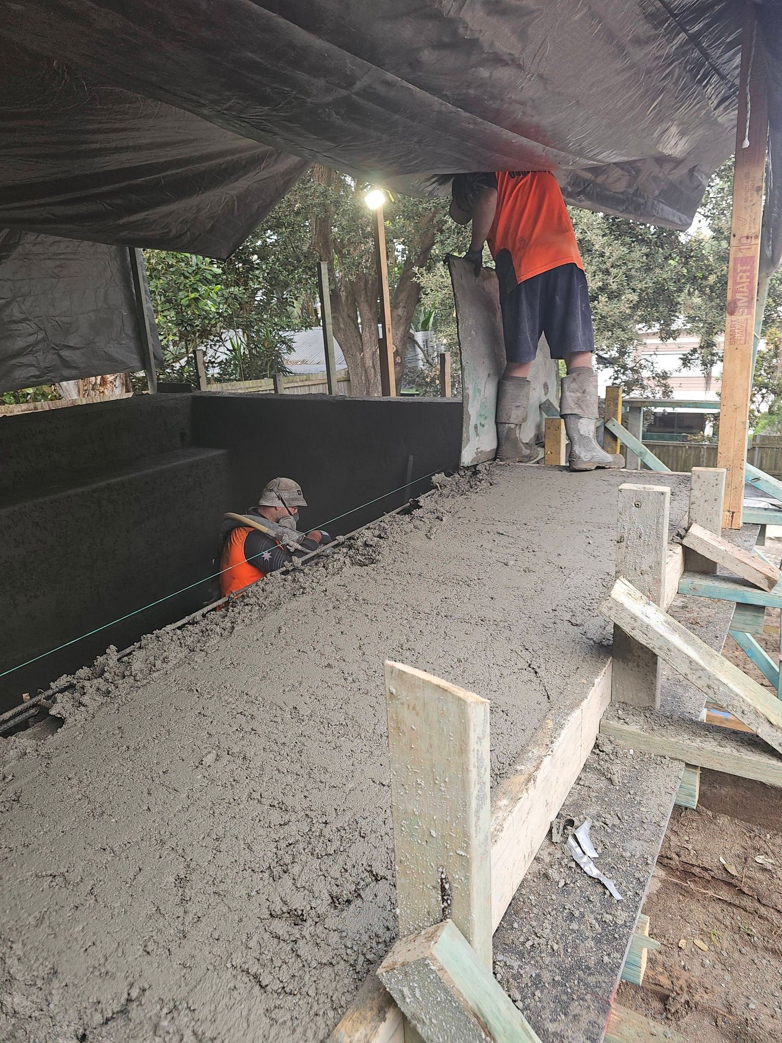 Workers smoothing fresh concrete on a sloped surface under a tarpaulin at a construction site