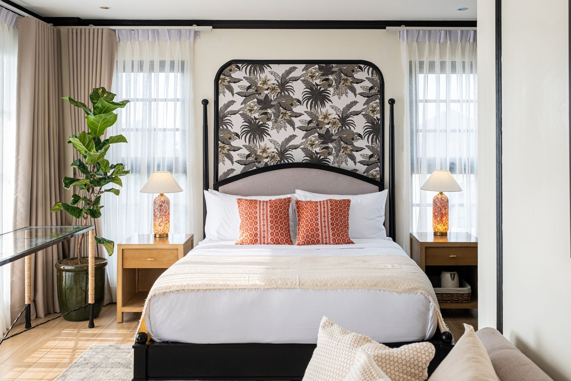 A bedroom with a bed featuring a floral headboard, two nightstands with lamps, a potted plant, and sheer curtains.