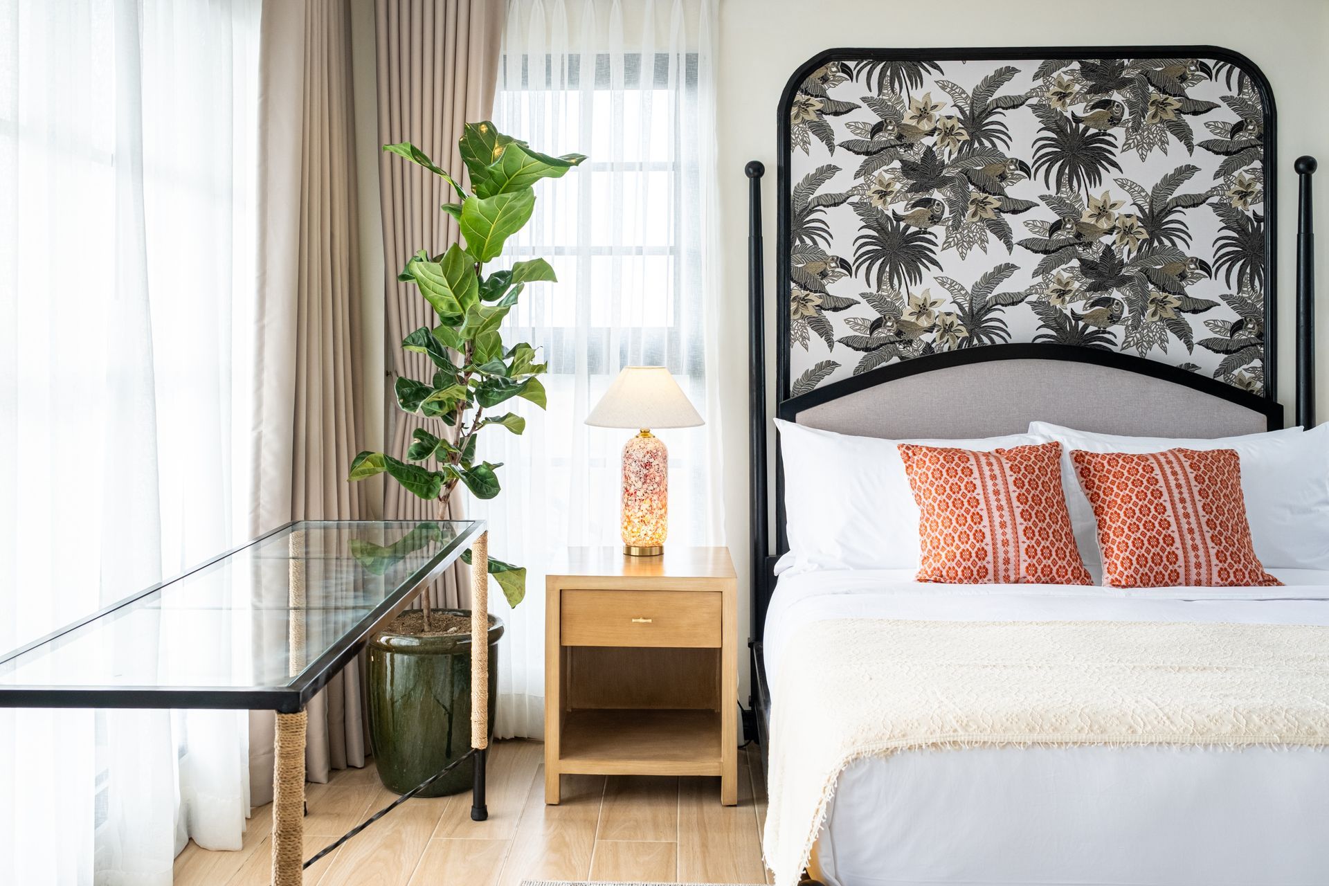 A bedroom with a bed featuring a botanical-patterned headboard, white linens, orange pillows, a nightstand, and a plant.