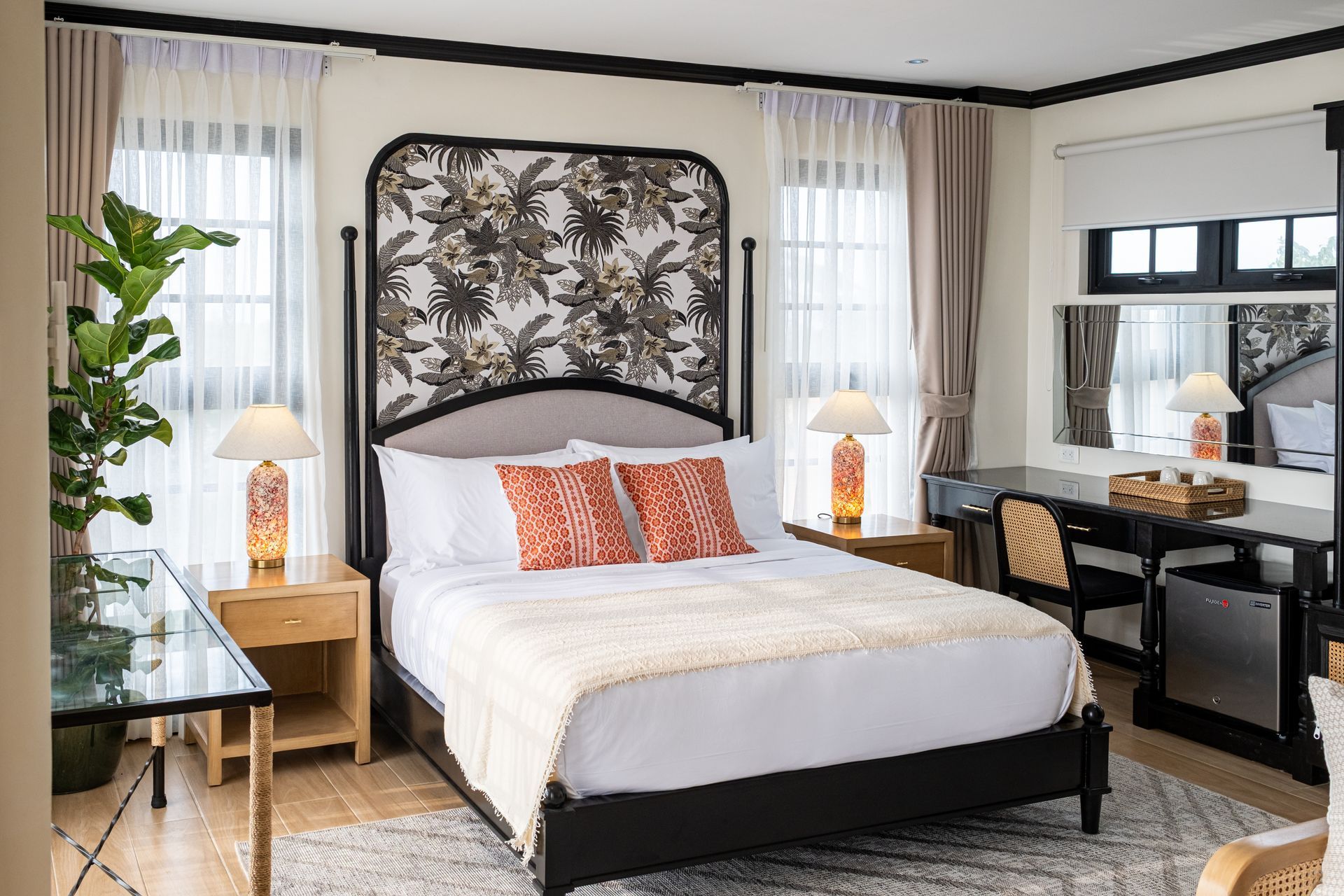 A hotel room with a queen bed featuring a floral headboard, two nightstands with lamps, and a glass-topped desk.
