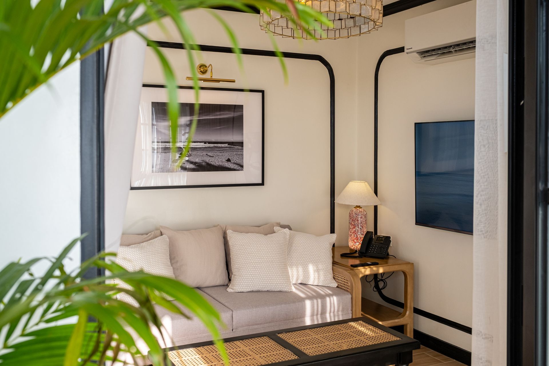 A cozy hotel seating nook with a gray sofa, throw pillows, a coffee table, and framed art, viewed behind a leafy plant.