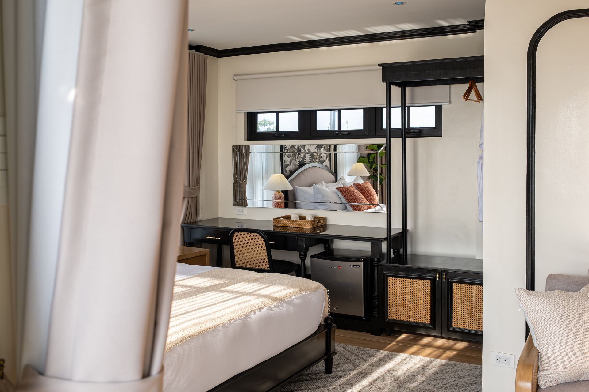 A modern hotel room with a bed, a desk, a mirror reflecting a bed, and rattan storage bins, bathed in soft, natural light.