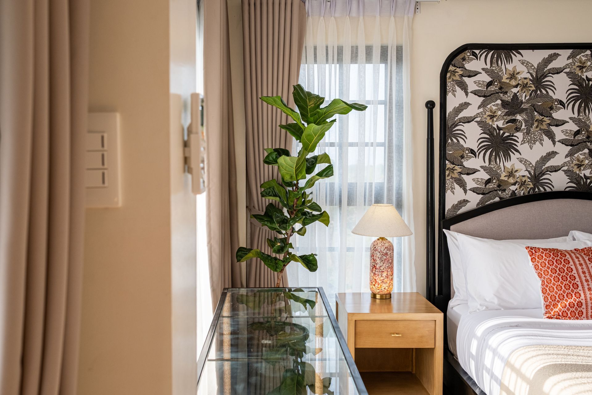 A bedside table with a lamp and a plant next to a bed with a tropical-patterned headboard, set near a curtained window.