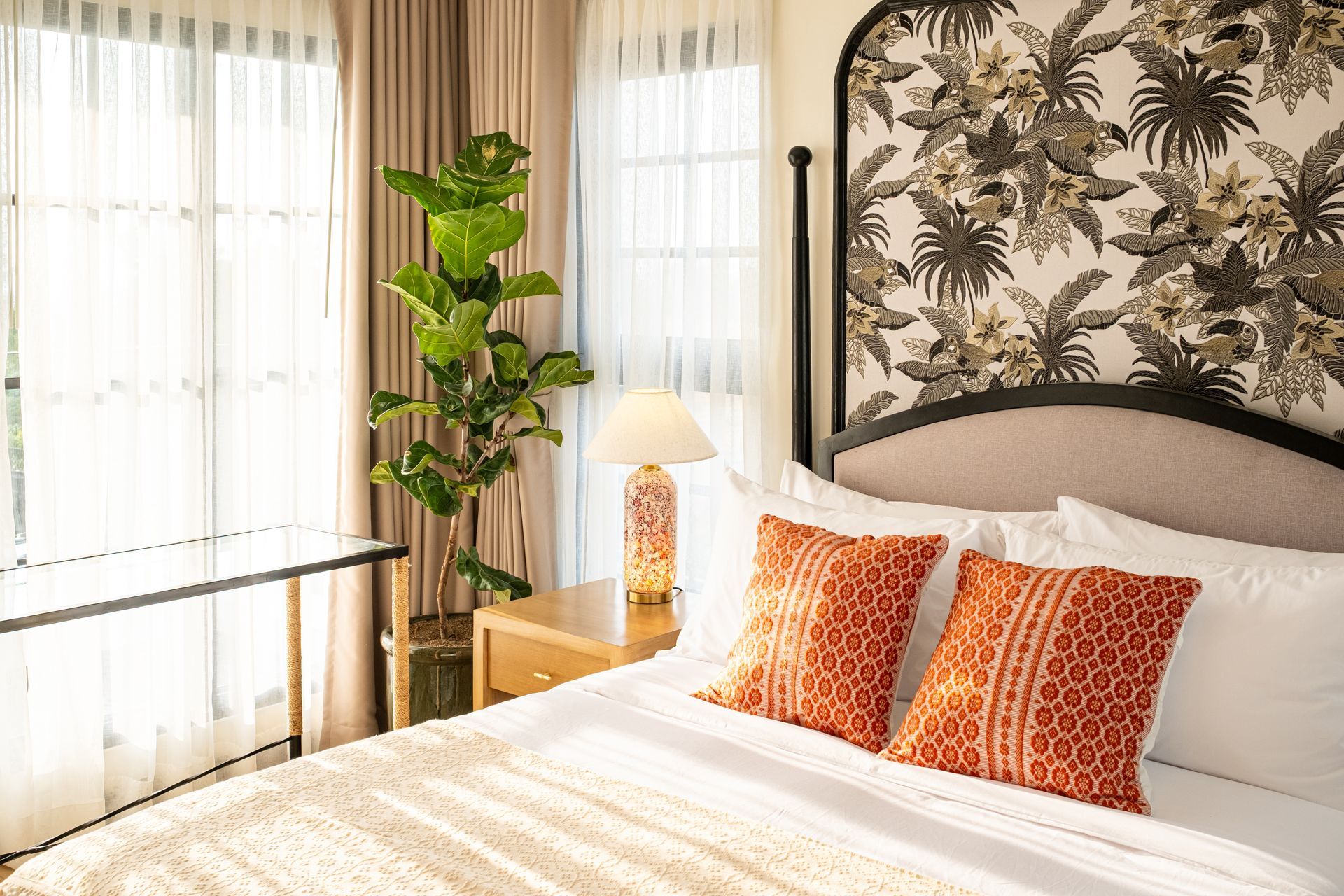 A bedroom with a patterned headboard, white bedding with orange accent pillows, a bedside lamp, and a tall potted plant.