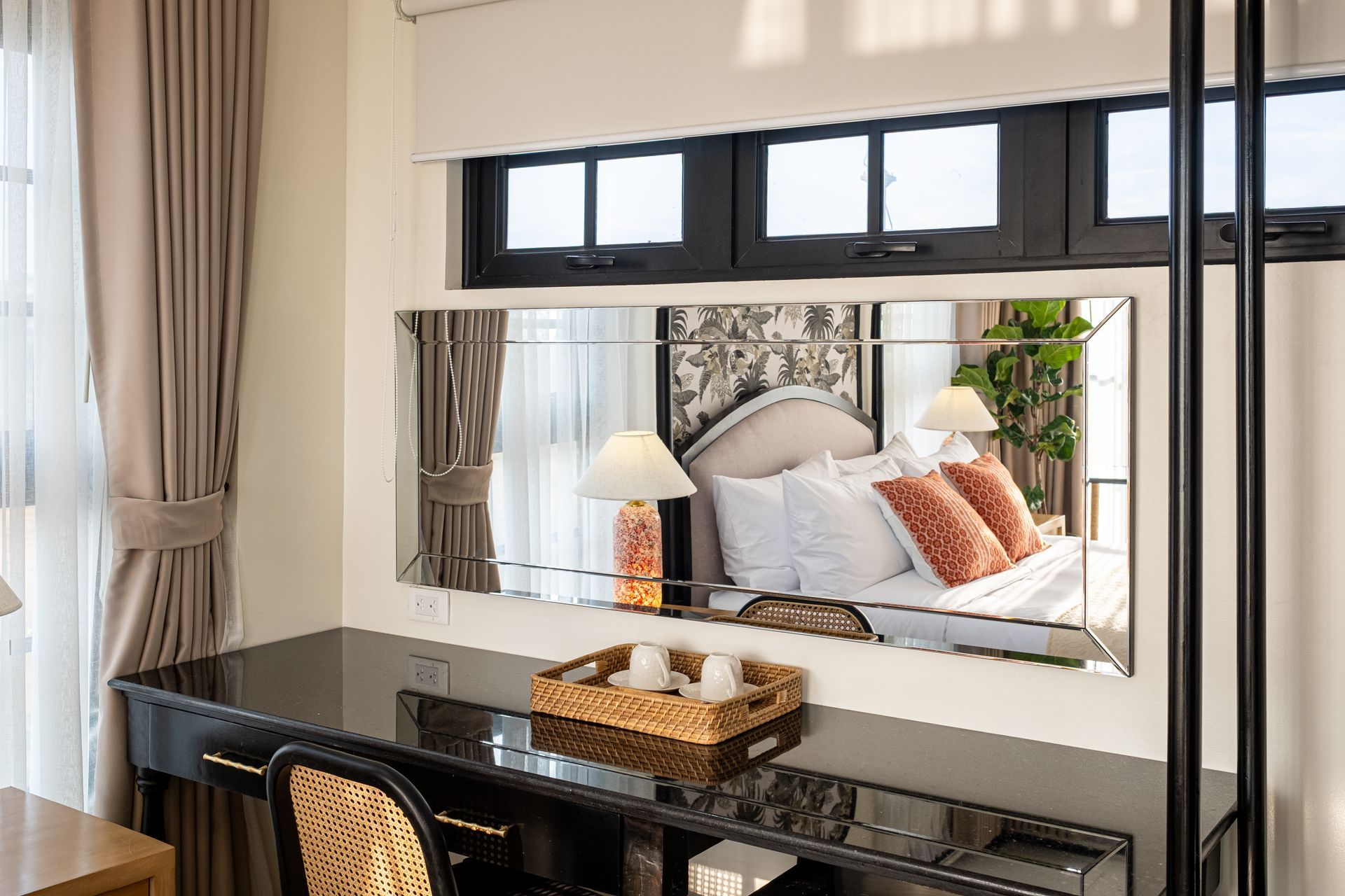 A desk with a wicker chair and tray, featuring a long, wall-mounted mirror reflecting a bed with decorative pillows.