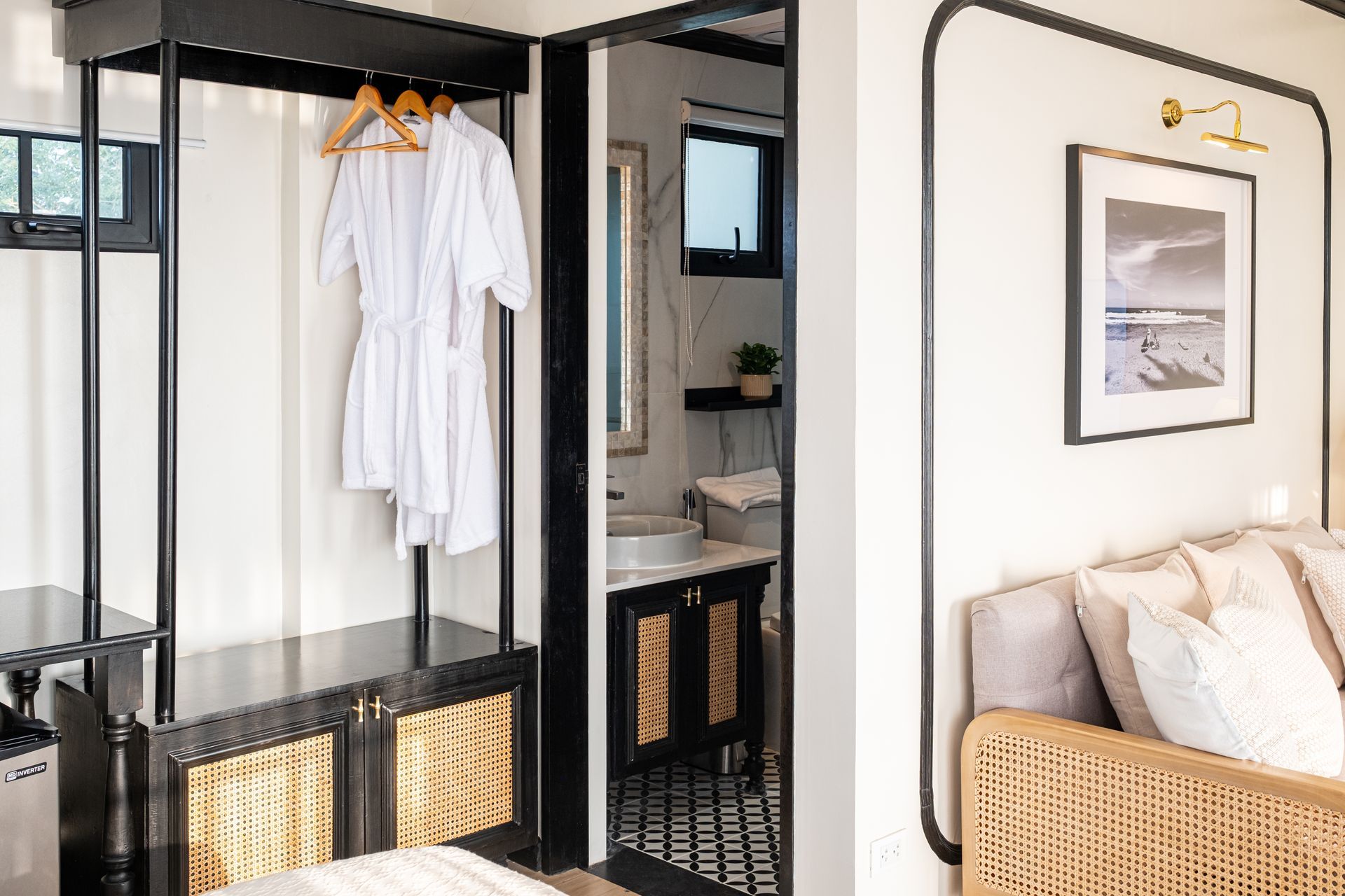 A bedroom with a hanging rack holding robes, a view into a bathroom with patterned tiles, and a wicker chair on the right.