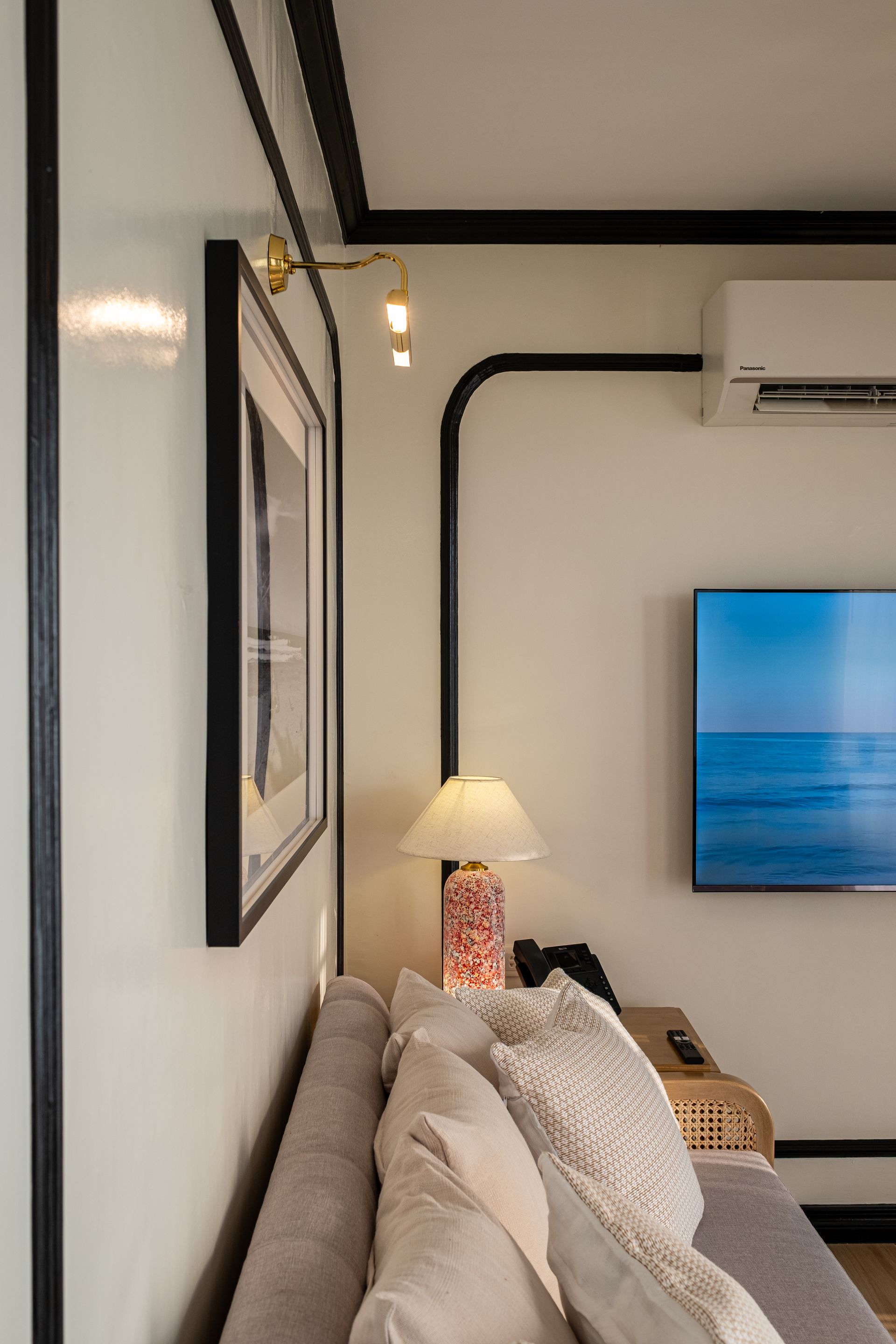 A sofa with patterned cushions sits in a hotel room with white walls, black trim, a wall-mounted TV, and bedside lamp.