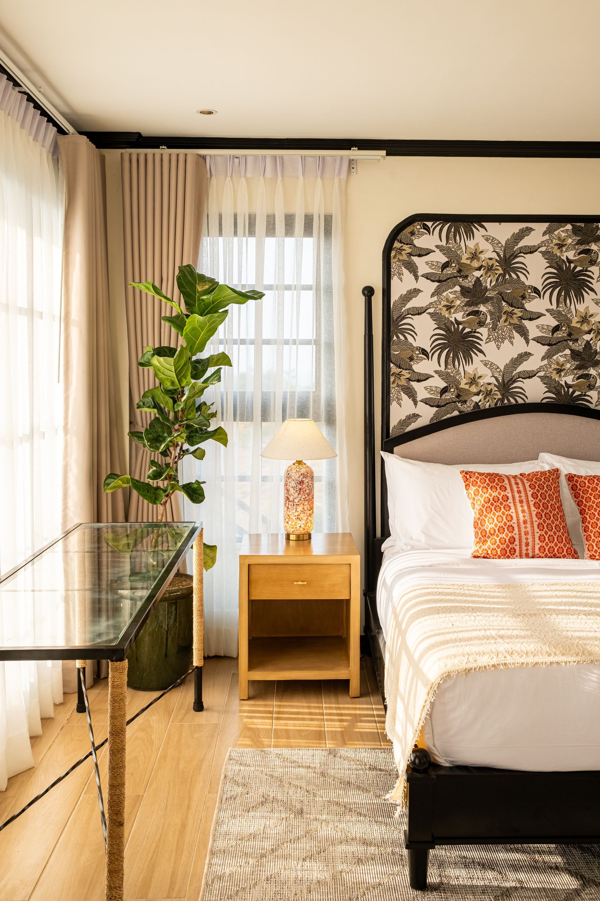 A bedroom featuring a bed with patterned headboard wallpaper, orange pillows, a glass desk, a floor plant, and a lamp.