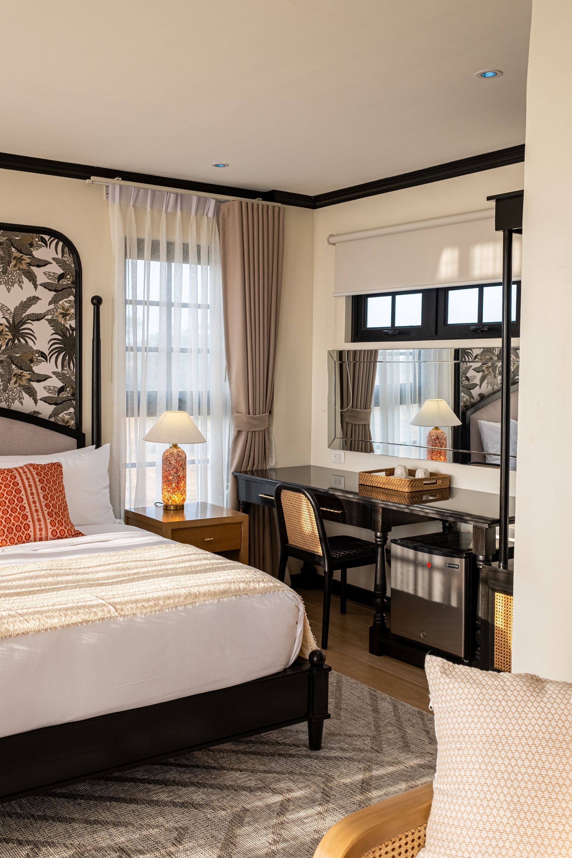 A hotel bedroom featuring a queen bed, a patterned rug, a wooden desk with a lamp, and sheer curtains on a window.