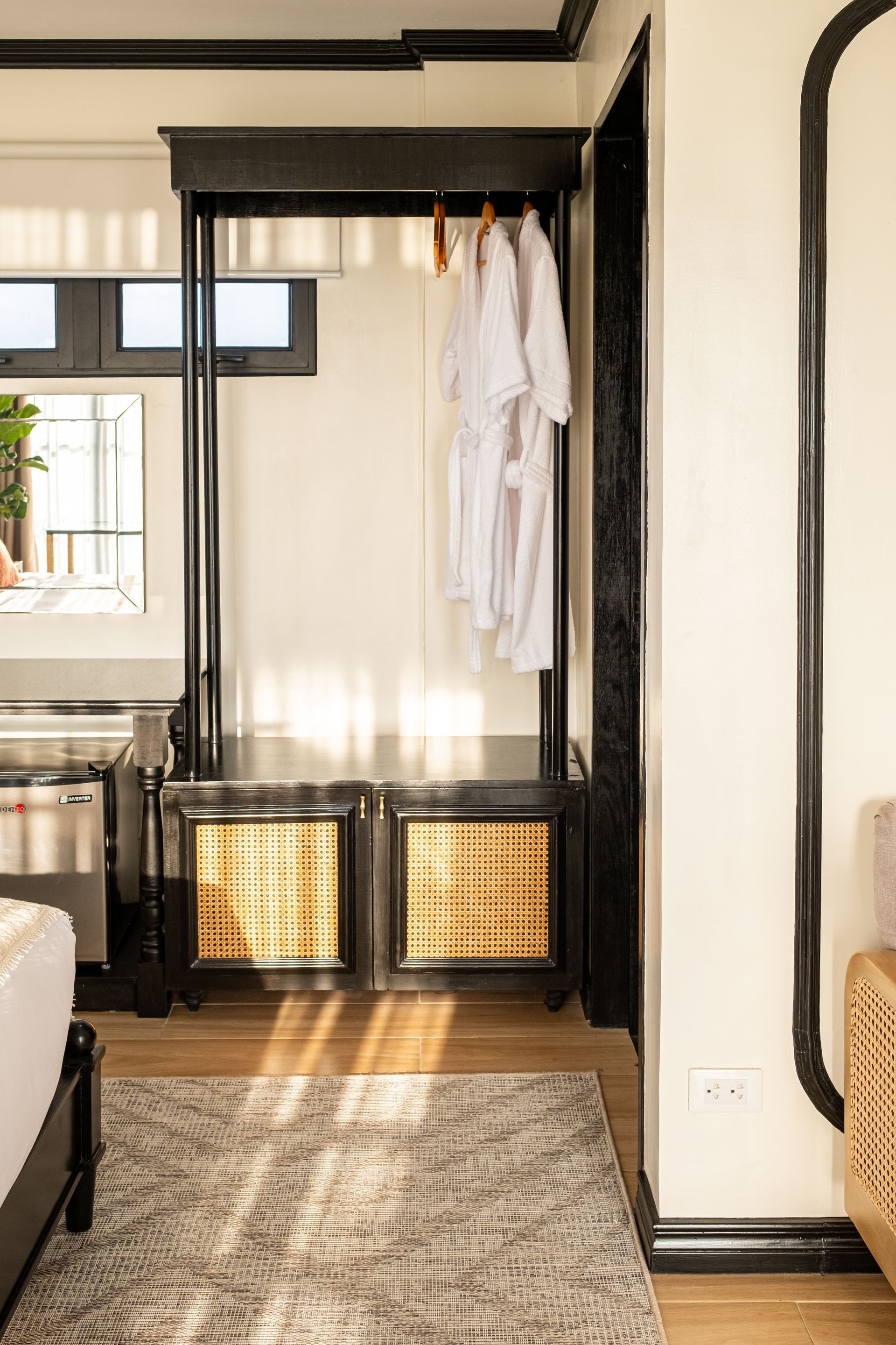 Black clothing rack with a cane-paneled base and white bathrobes hanging inside, set against a cream-colored wall.
