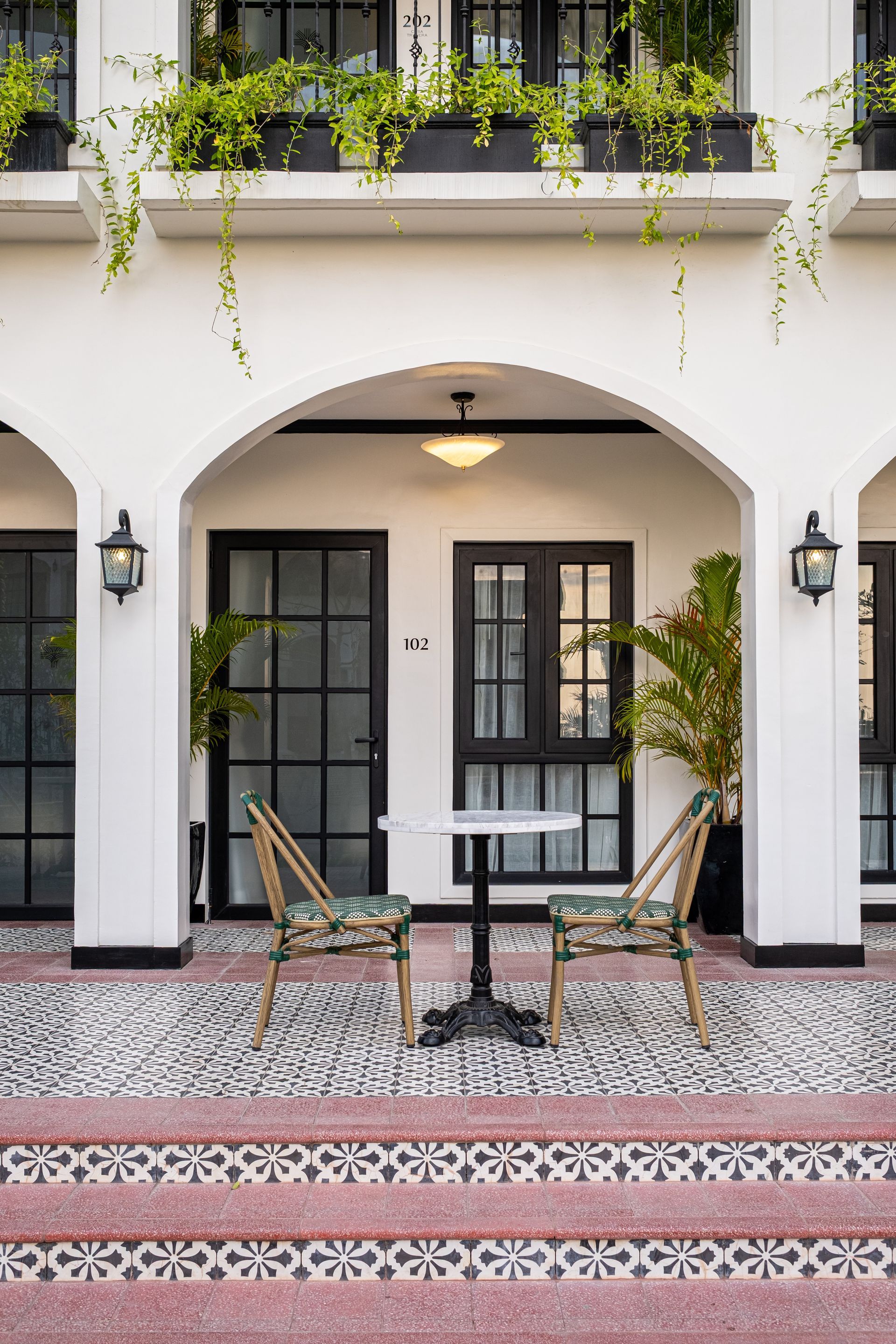 A white building with arched entryways, a small bistro table with two chairs, and lush hanging plants on the balcony.