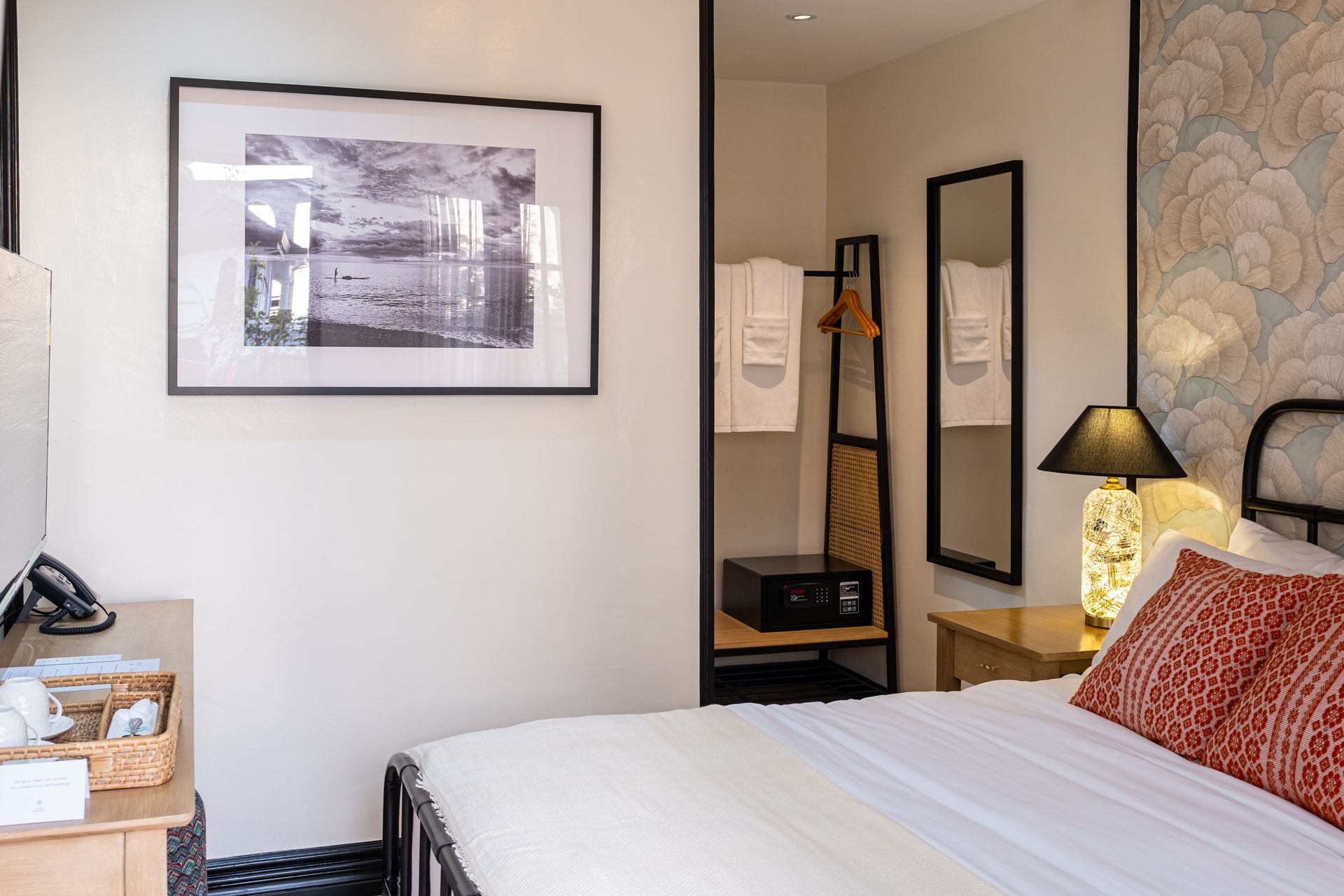 A cozy bedroom with white walls, patterned wallpaper, a black bed frame, a framed landscape print, and a bedside lamp.