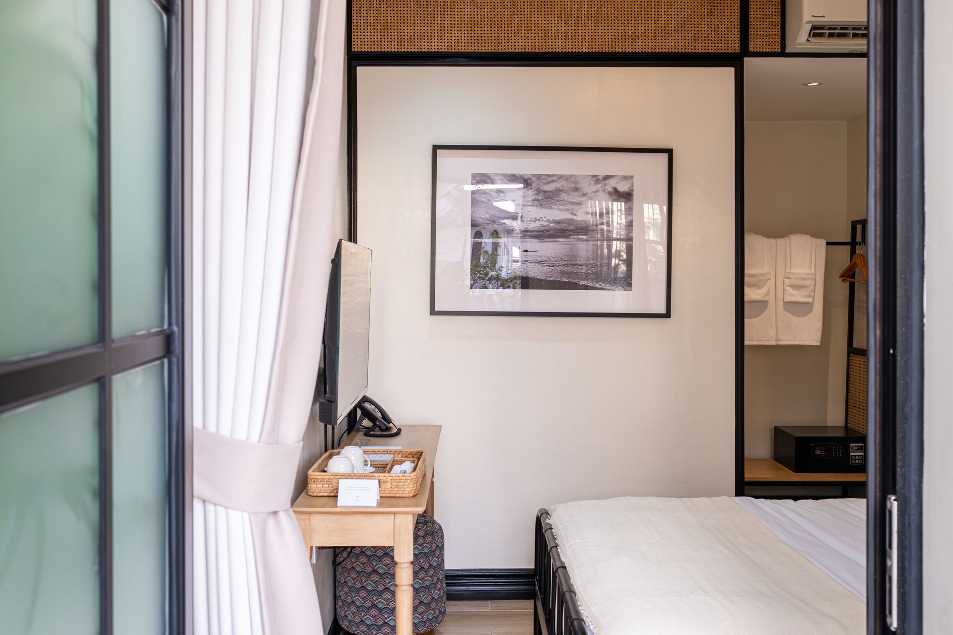 A view through a door into a hotel room featuring a small desk with a TV, a framed wall print, and a bed corner.