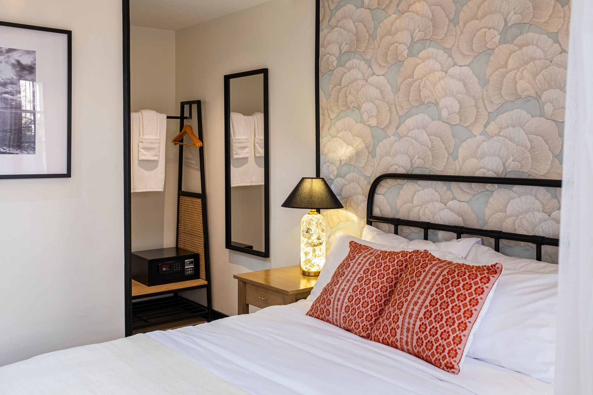 A hotel room with a bed featuring orange patterned pillows against floral wallpaper, a bedside lamp, and a small closet.