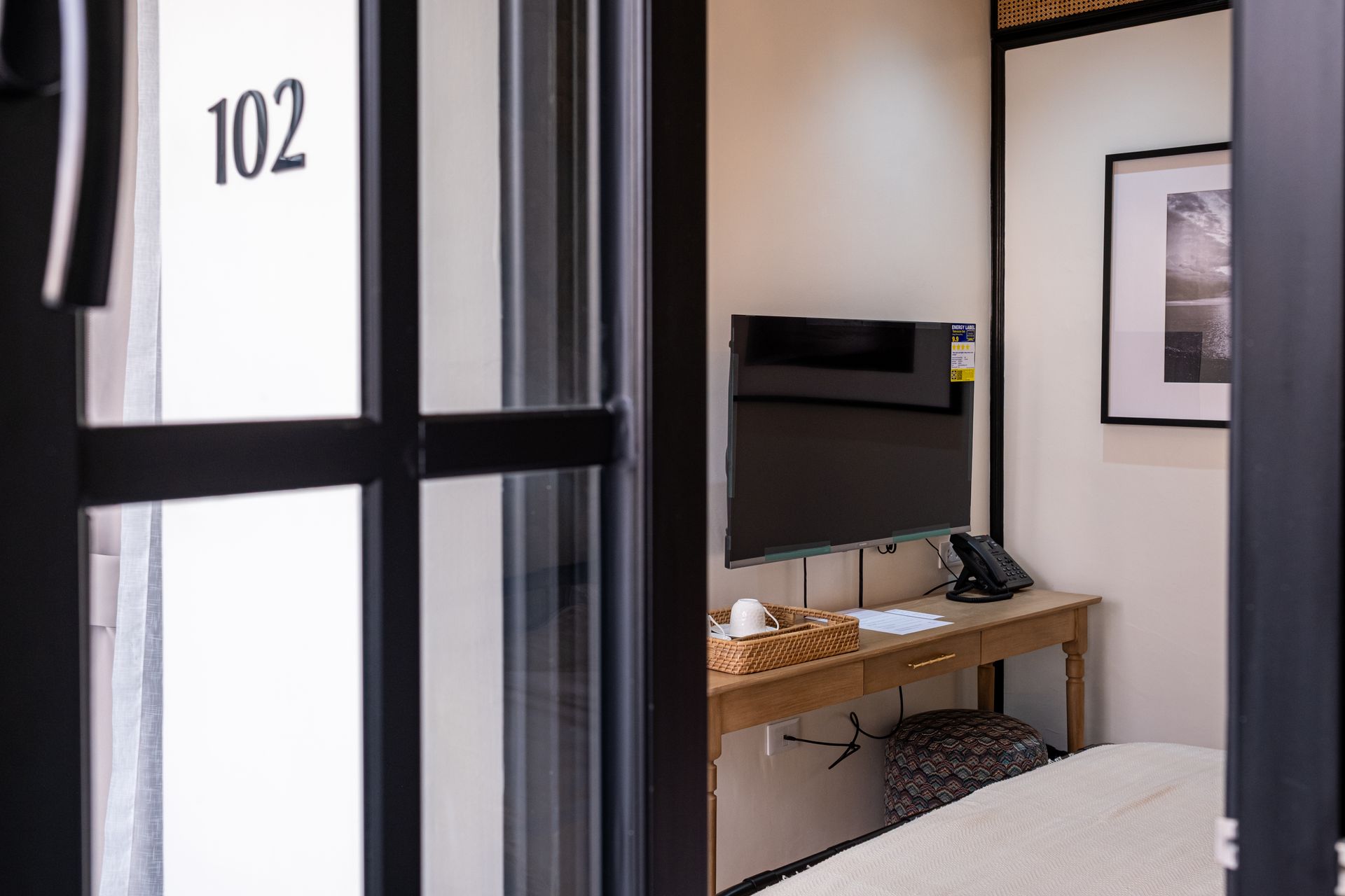 View of a hotel room numbered 102, featuring a flat-screen TV on a desk with a wicker basket and a partial bed view.
